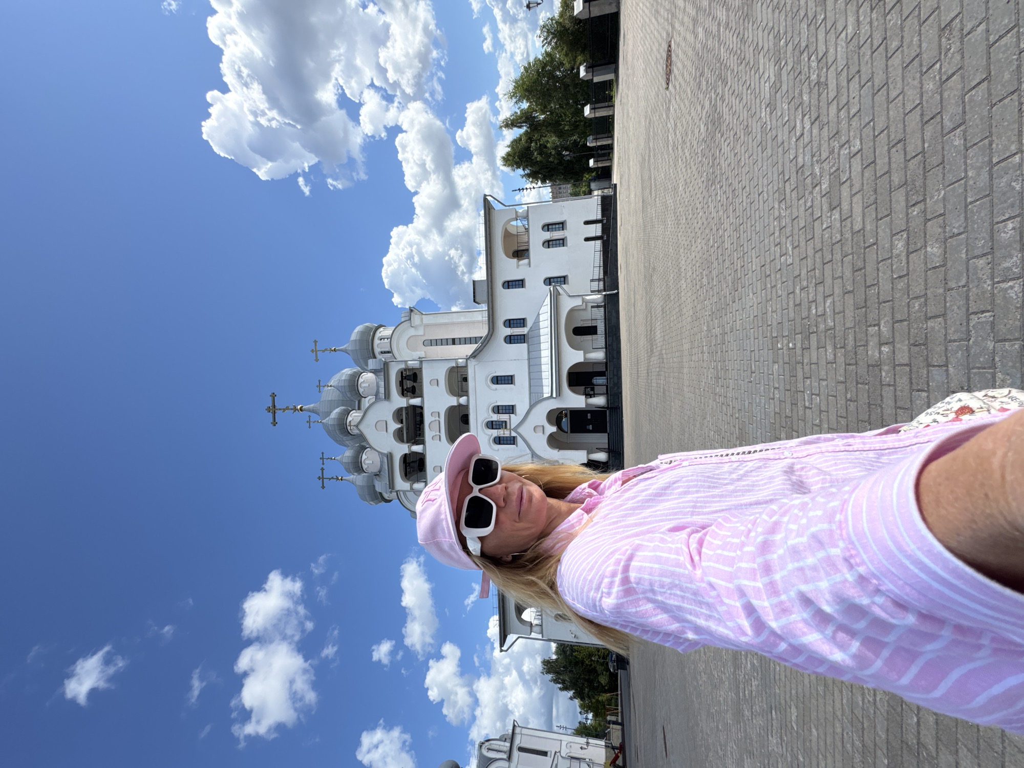 A blonde woman with sunglasses and a pink striped shirt and cap stands in front of a white church with silver domes under a blue sky with clouds. The ground is paved with gray bricks. The church appears to be the Church of the Icon of the Mother of God "The Joy of All Who Sorrow" on Vasilievsky Island in St. Petersburg, Russia.