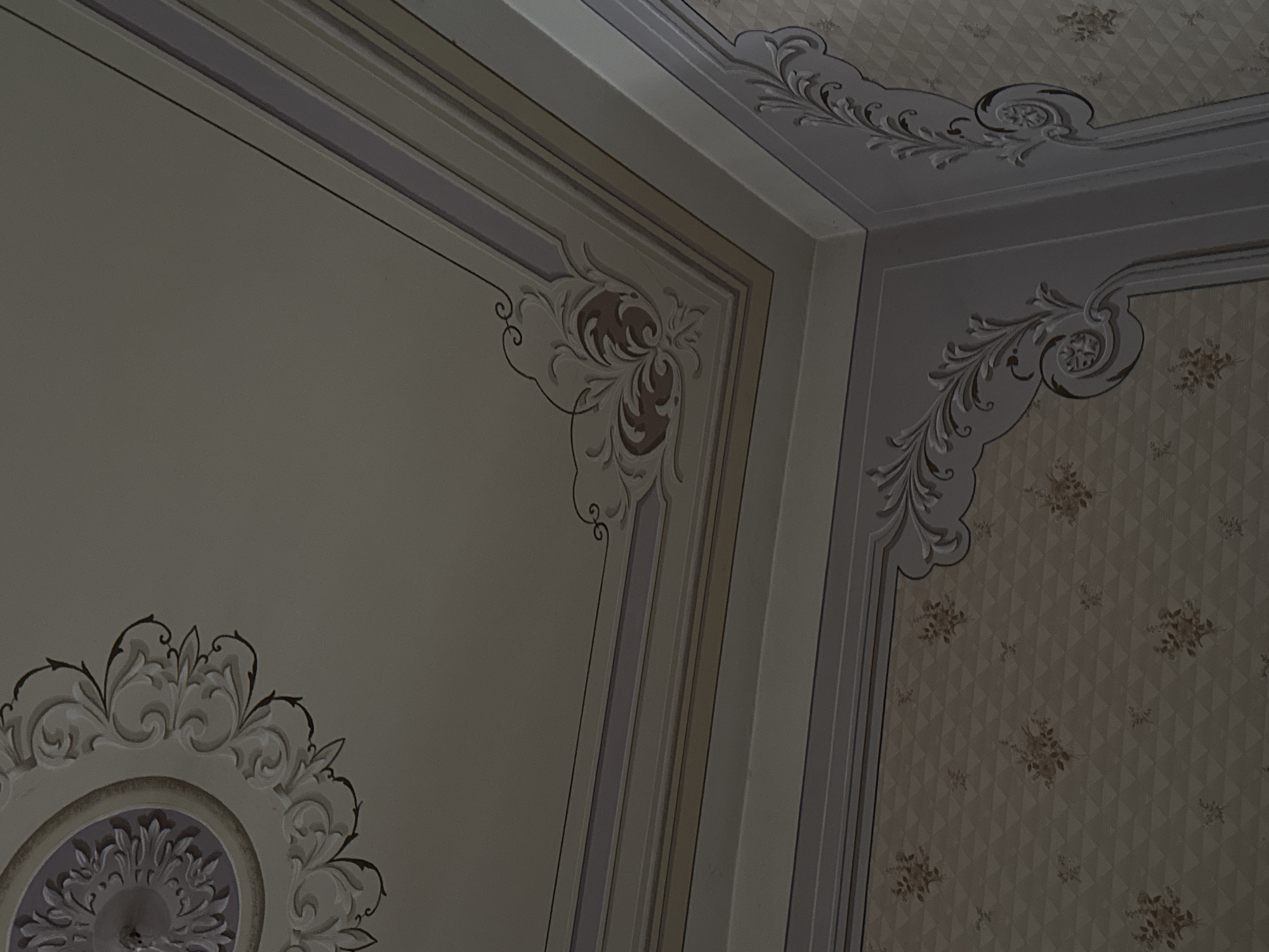 The image shows a corner of an ornate interior, likely the ceiling and upper wall area. The location is Tbilisi, Georgia. The ceiling and wall feature decorative molding and intricate floral or leaf-like patterns. One section of the wall shows a patterned wallpaper with a diamond or triangular grid and small floral designs. The color palette is muted, consisting of off-whites, creams, and subtle browns. The scene appears to be an interior shot of a historic building, possibly a palace or a grand residence.