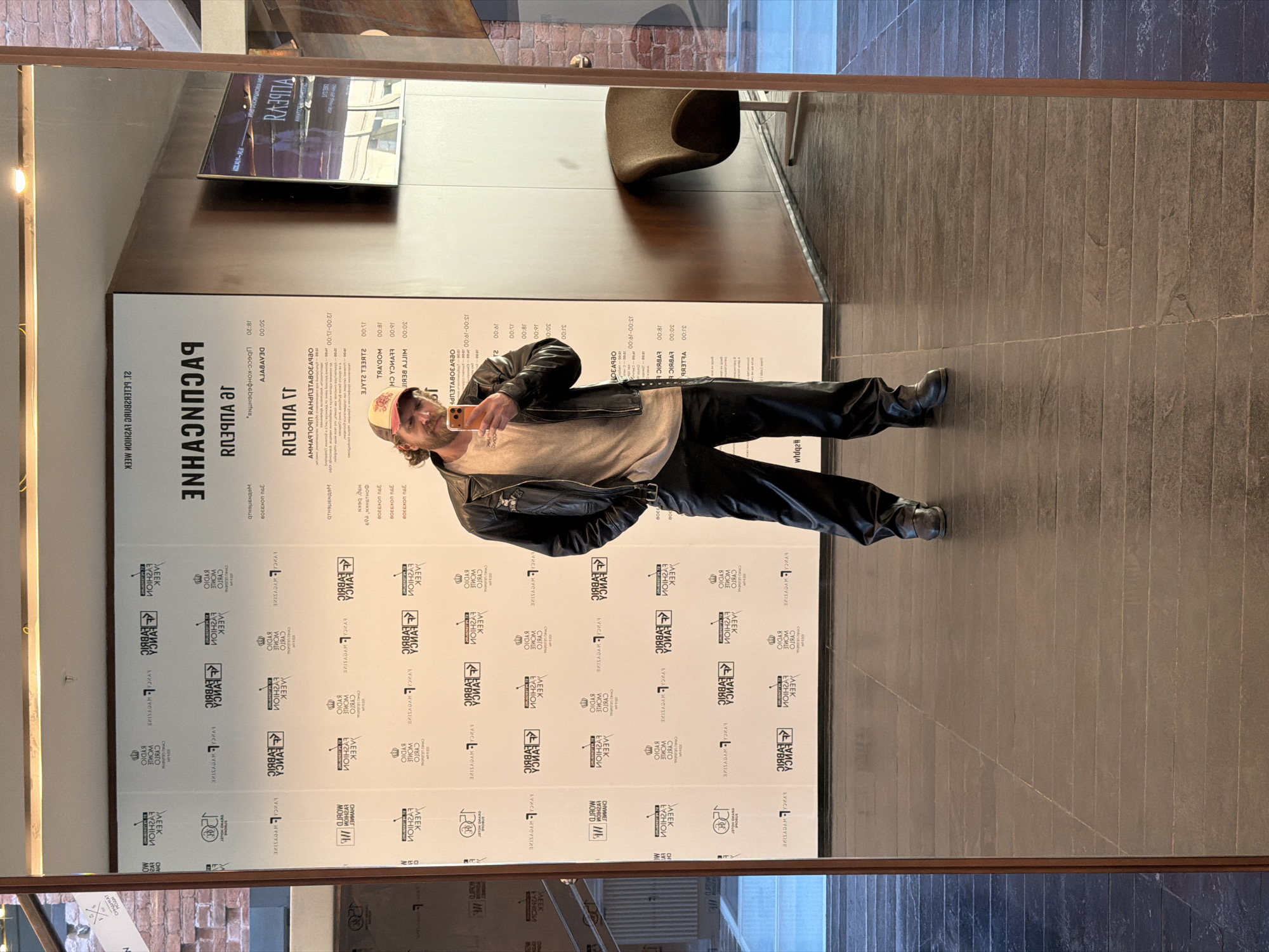 An indoor, vertical mirror selfie captures a man posing in front of a large event schedule poster. The man, likely the photographer, is seen from the chest down, wearing a black leather jacket over a light-colored, ribbed long-sleeve shirt, dark pants, and black boots. He has a beard and a baseball cap worn backward, and holds a smartphone in his right hand, seemingly to take the photo. A slight smirk or smile is visible on his face.
The reflection shows a spacious interior with dark tiled flooring and a dark, possibly wooden, wall. A dark, modern-looking chair is also visible in the background. The primary background element is a large white banner or poster, featuring text predominantly in Russian Cyrillic, indicating the event's location in Saint Petersburg, Russia.
The prominent text on the banner reads "РАСПИСАНИЕ" (SCHEDULE) and "21 АПРЕЛЯ" (April 21), suggesting a one-day event or the schedule for that specific date. A list of times and associated activities or names is presented, including "ПОКАЗЫ" (Shows/screenings), "ЛЕКЦИИ" (Lectures), "Цвесс-конференция" (Press conference), and names like "ВЕЛЛЕР" (Weller) and "МИГГИ БЕРГ" (Miggy Berg). Repeated logos and text towards the bottom of the banner feature phrases like "НЕДЕЛЯ МОДЫ" (Fashion Week) and "КУБРИК КИНО," "КУБРИК ВЫСТАВКА," "КУБРИК КОНЦЕРТ" (Kubrik Cinema, Kubrik Exhibition, Kubrik Concert), implying a multi-faceted cultural or fashion event hosted by a venue named Kubrik. The reflection in the mirror also shows a distant view of what appears to be an exterior building or street scene, slightly blurred.