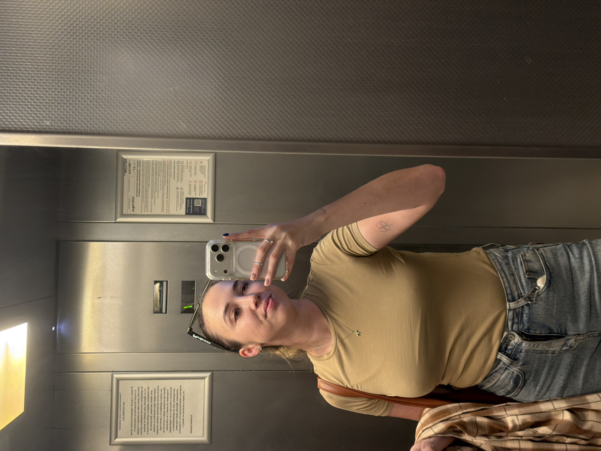 The image shows a woman taking a selfie in an elevator. She is wearing a tan t-shirt, blue jeans, and a brown shoulder bag. She has glasses on her head and a necklace. In her right hand, she holds a smartphone, and on her left arm, she has a tattoo of a four-leaf clover. The elevator walls are metallic with a textured pattern. There are two framed notices on the left wall of the elevator. Based on the presence of Cyrillic text on the notices, the location is likely Rostov-on-Don, Russia.