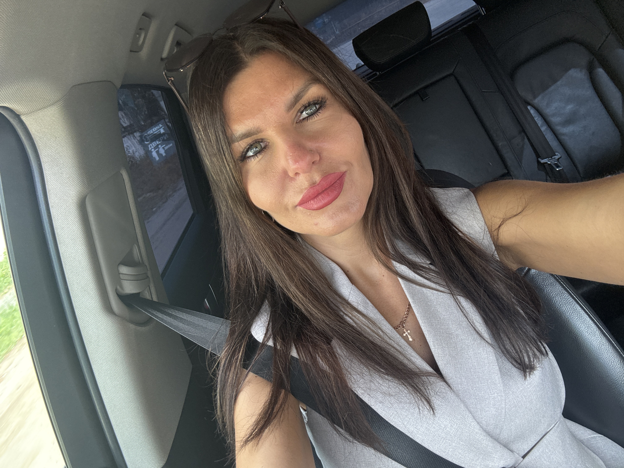 The image shows a woman with long, dark brown hair and green eyes, wearing a light-colored blazer and a cross necklace, sitting in the driver's seat of a car. She is taking a selfie, with her face filling most of the frame. The interior of the car, including the black leather seats and a gray seatbelt, is visible. Through the window, a blurred outdoor scene is visible, suggesting the car is in motion. The location is identified as Chaikovsky, Russia, but no specific landmarks or details of the city are apparent in the image.
