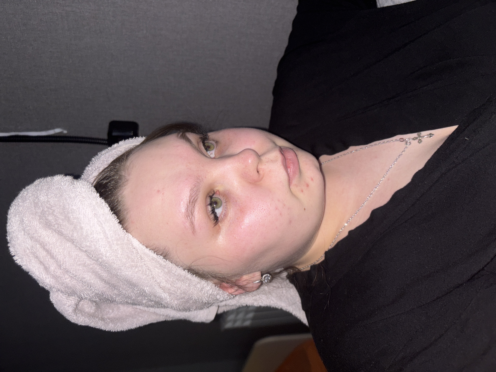 The image presents a young adult female indoors in Chaikovsky, Russia, captured in a chest-up portrait. Her hair is wrapped in a light pink terry cloth towel. Her face shows some redness and minor blemishes on the cheeks, chin, and forehead. She has green eyes with notable eyelashes, directed towards the viewer's left. Her left ear is visible, adorned with a silver-colored hoop earring featuring a circular cluster of clear stones. A thin silver-colored chain necklace with a small cross pendant rests on her chest, over a black, ribbed V-neck garment. The background consists predominantly of a dark grey textured wall. A dark rectangular object with an attached cable is visible on the upper right side of the wall. Hints of a light-colored object and an orange surface are partially visible in the blurred background on the left.