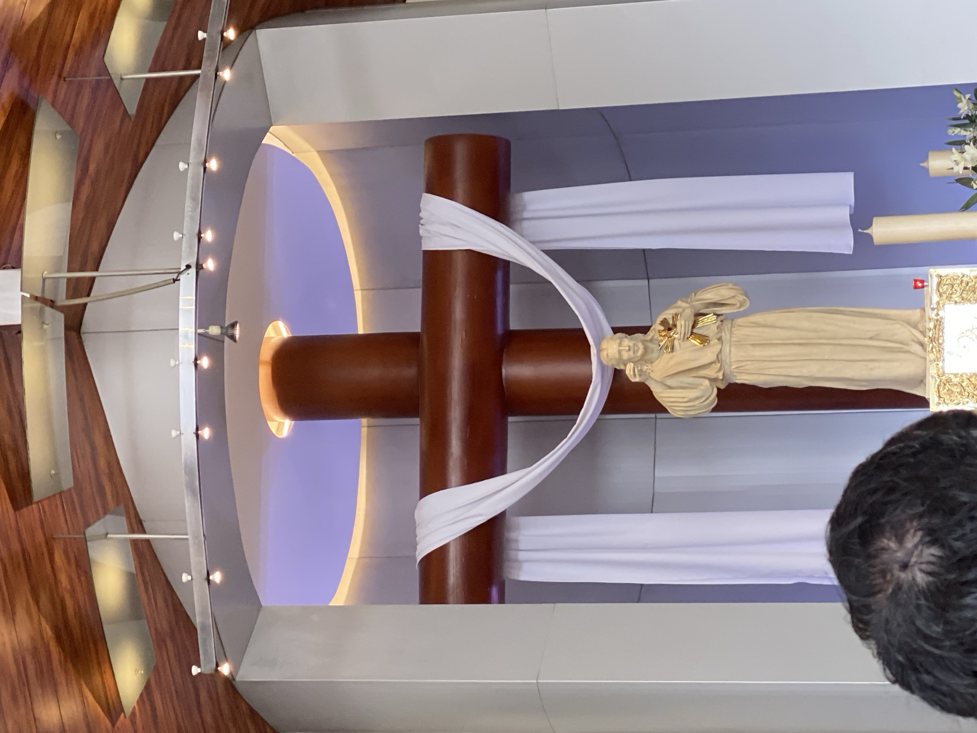 The image depicts the interior of a church in San Bernardino Tlaxcalancingo, Mexico. A prominent crucifix with a draped white cloth is centrally positioned. To the right of the cross is a statue of a saint with two candles. The background features an archway with illuminated edges and a series of lights along the perimeter. The structure is primarily constructed of wood and light-colored materials. In the lower right corner, a person's hair is partially visible.