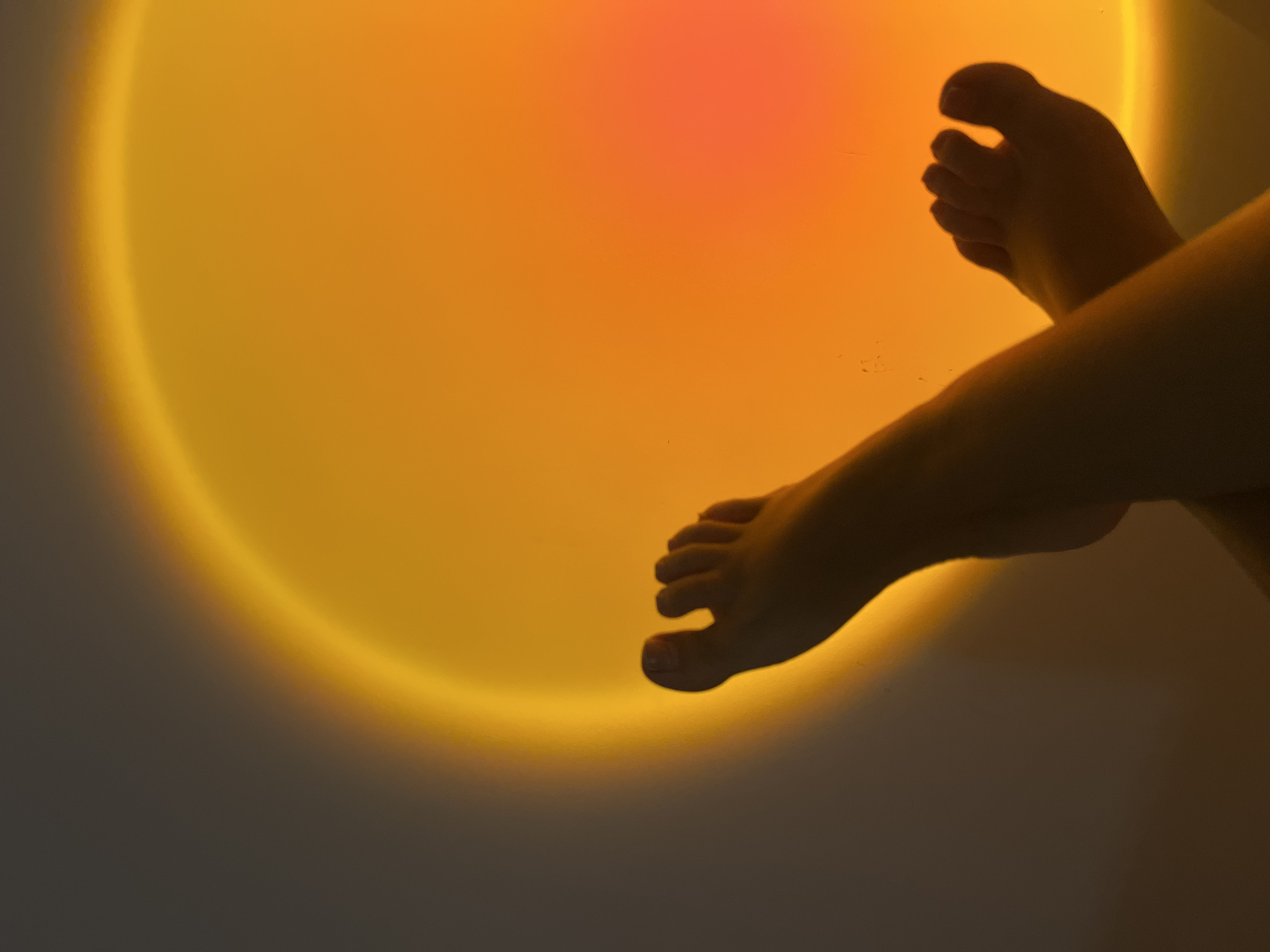 The scene is located indoors in Kiryat Bialik, Israel. The primary subjects are two human feet, silhouetted against a large, circular light projection on a vertical surface, likely a wall. The light displays a gradient, with an outer ring of yellow-orange transitioning to a central area of reddish-orange. The feet are bare and positioned in a crossed arrangement, with the left foot elevated above the right. Toenail polish is visible on the digits of the upper left foot. The environment outside the light projection appears dark grey to black, indicating low ambient lighting. The perspective is upward, capturing the feet in the lower half of the frame with the illuminated wall dominating the background.