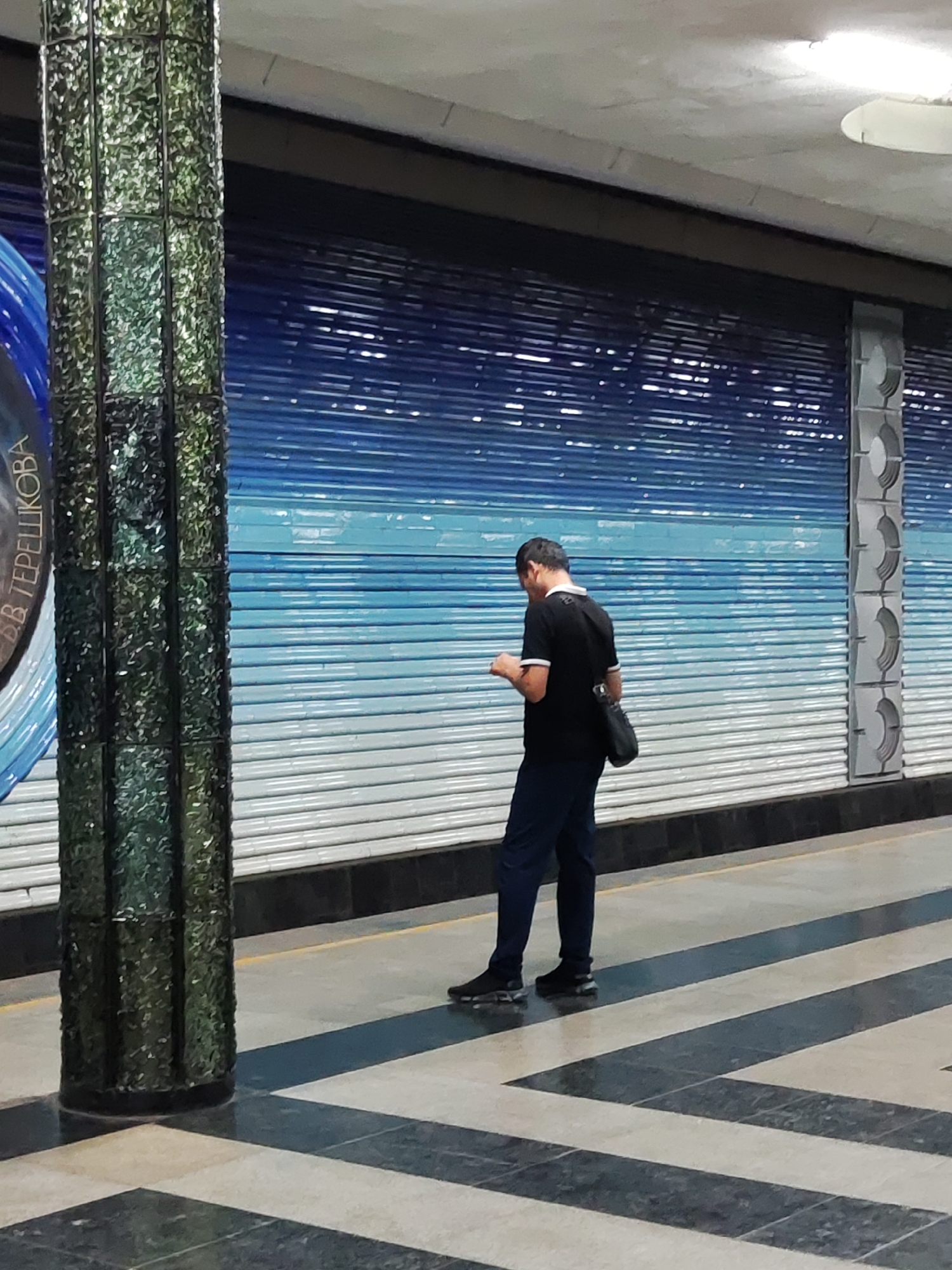 A man in a black shirt and dark pants stands looking at his phone on a subway platform in Tashkent, Uzbekistan. The platform has a checkered floor and a wall with a blue and white striped design. A green tiled pillar is in the foreground.