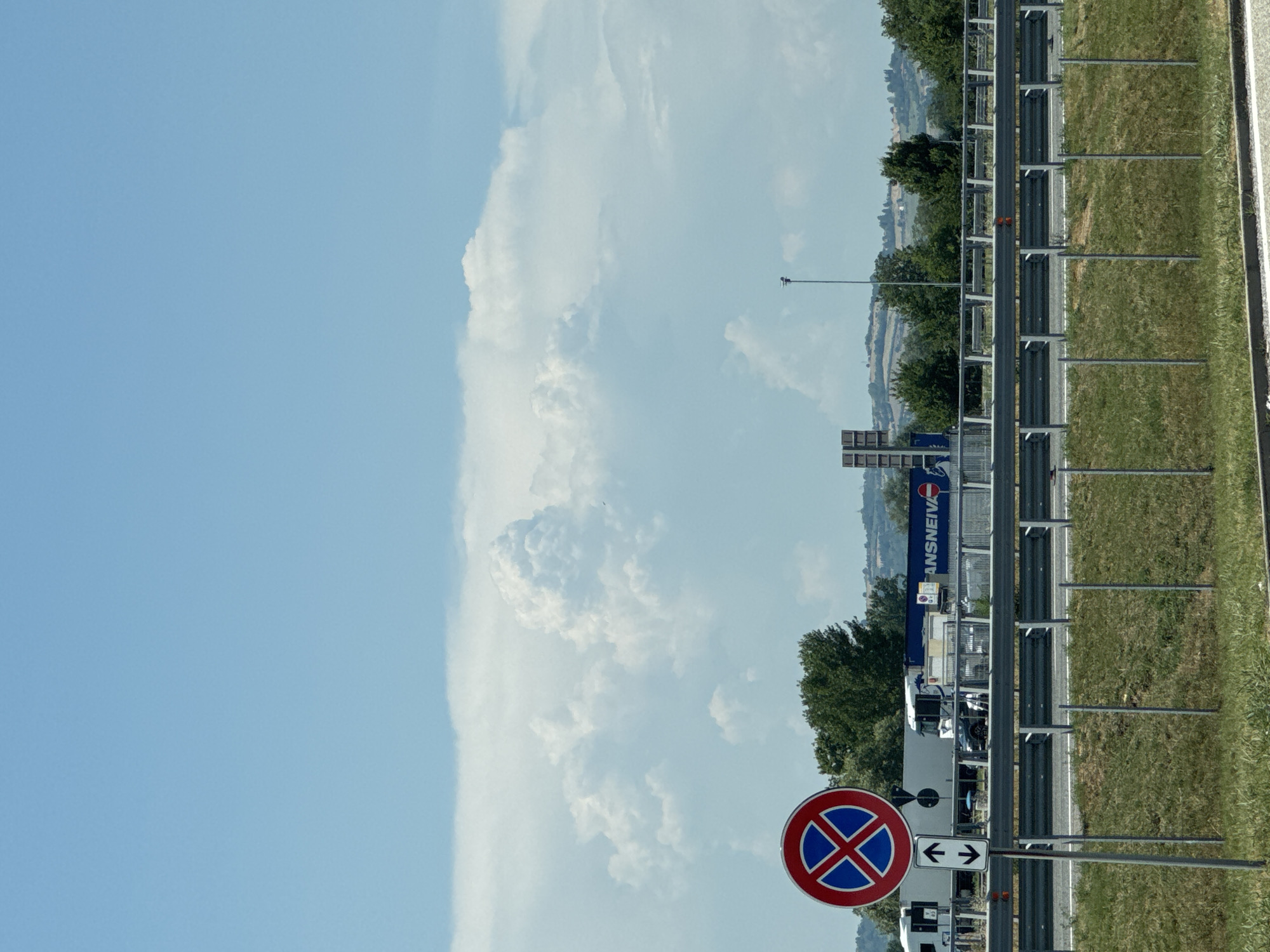 The image shows a roadside scene on a sunny day, likely in or near Occimiano, Italy, given the context of its metadata. In the foreground, a grassy embankment is bordered by a metal guardrail. To the left, a traffic sign with a red circle and a blue "X" indicating no stopping is prominently displayed, accompanied by a smaller sign with vertical arrows pointing up and down.
Behind the guardrail, a road is visible with several vehicles. A large white truck with a blue stripe and the partial word "ANSNEIVA" on its side is parked or stopped. A smaller white vehicle is positioned behind it. Further in the background, a line of trees forms a natural barrier, with rolling hills and some buildings visible in the distance. The sky is a clear blue, with large, puffy white clouds dominating the upper portion of the frame. A single street lamp stands to the right of the frame in the mid-ground. The overall impression is one of a typical highway or main road setting in a rural or semi-rural Italian landscape.