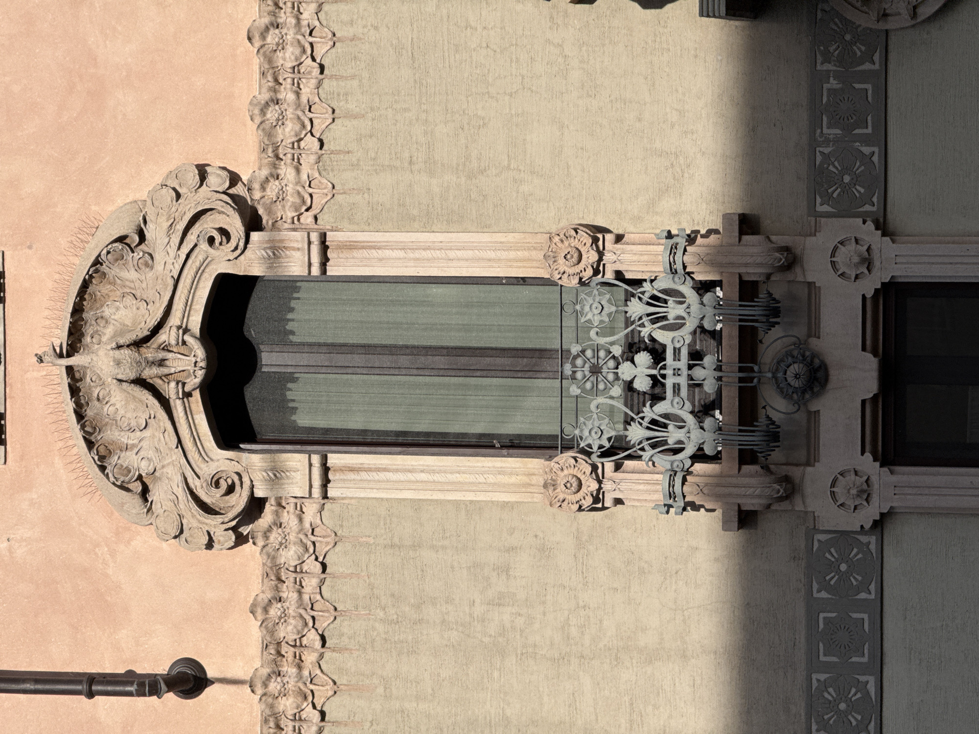 The image shows an ornate window on a building facade. The window has two panes of glass and is surrounded by elaborate stone carvings. The carvings include floral patterns, geometric shapes, and a sculpted bird. A metal grill with a floral design is partially covering the window. The building's facade is painted in two different colors: a light peach and a light green. There is a pipe mounted on the wall, next to the window. The photo shows architectural details and likely could be associated with a historical building in Milan, Italy, known for its architectural history.