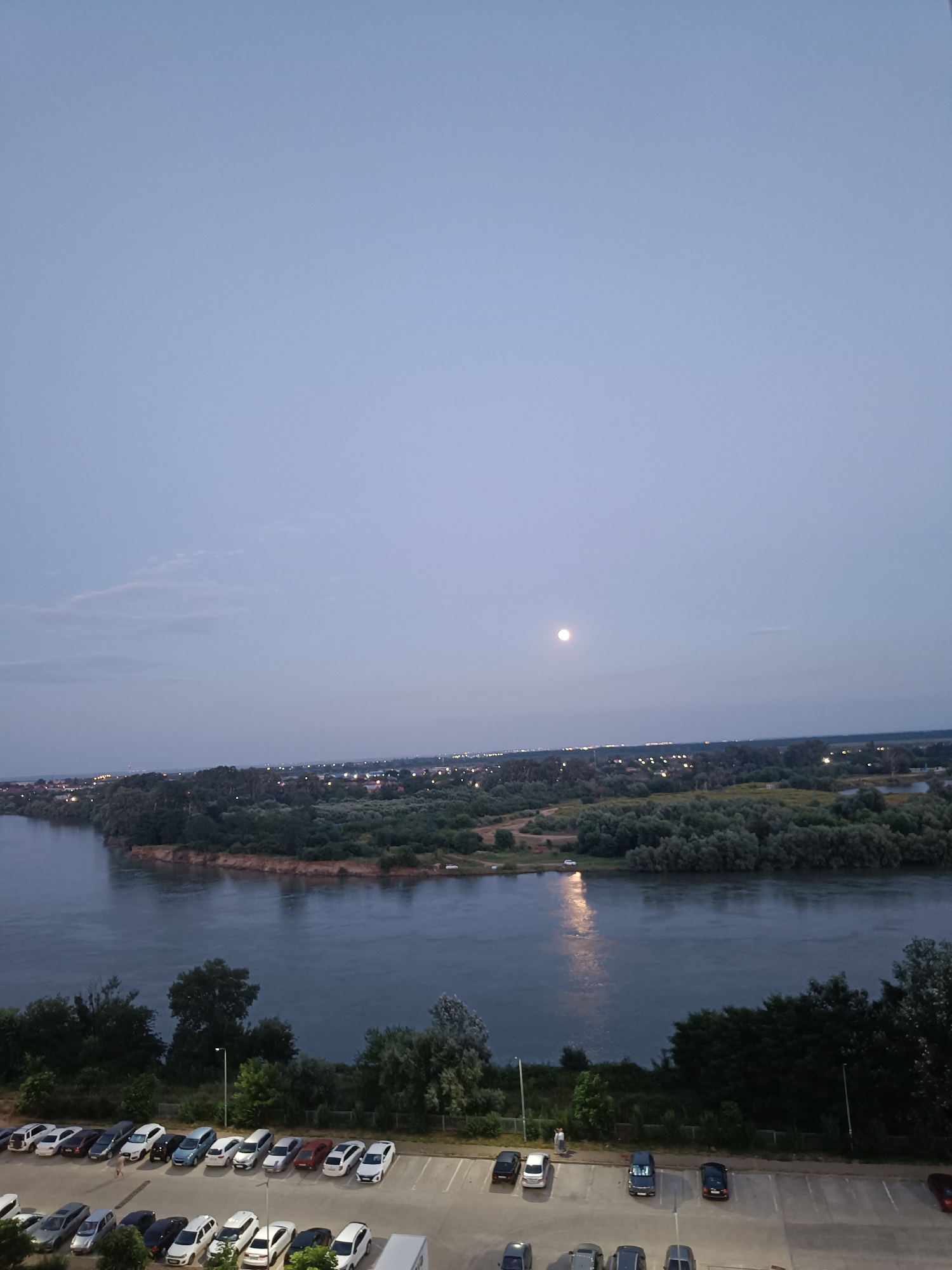 This image captures a panoramic view of the Kuban River in Krasnodar, Russia, during dusk. The scene is dominated by the wide, calm river reflecting the twilight sky and the lights of the city in the distance. The far bank of the river is lined with dense green trees and a settlement with illuminated buildings, creating a soft glow against the darkening horizon. Above the city, a full moon shines brightly in the pale blue sky. In the foreground, a parking lot is visible, filled with a variety of cars parked in neat rows. A single person is standing in the parking lot, seemingly observing the scenery. The overall atmosphere is tranquil and serene, showcasing the natural beauty of the river juxtaposed with urban elements.