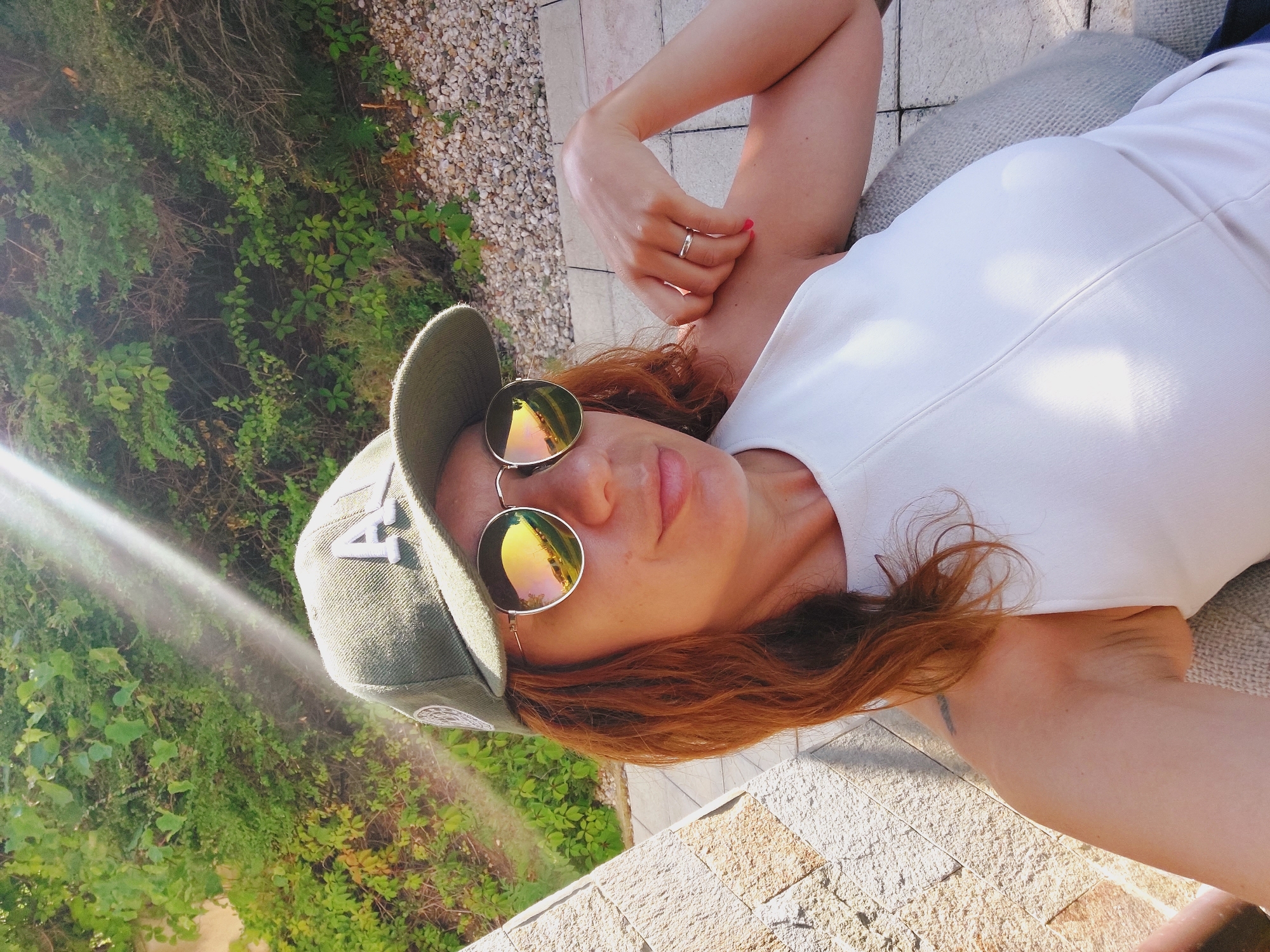 A woman with auburn hair, wearing a white sleeveless top, a green baseball cap with an "A" logo, and round, reflective sunglasses, is taking a selfie outdoors. She is seated, with her left arm resting on her lap and her right arm bent, her hand near her shoulder. The background shows lush green foliage, a stone wall to her left, and a gravel area with paving stones. Sunlight streams down from the upper left, creating lens flare. The scene appears to be a residential outdoor space or a garden in Sozopol, Bulgaria.