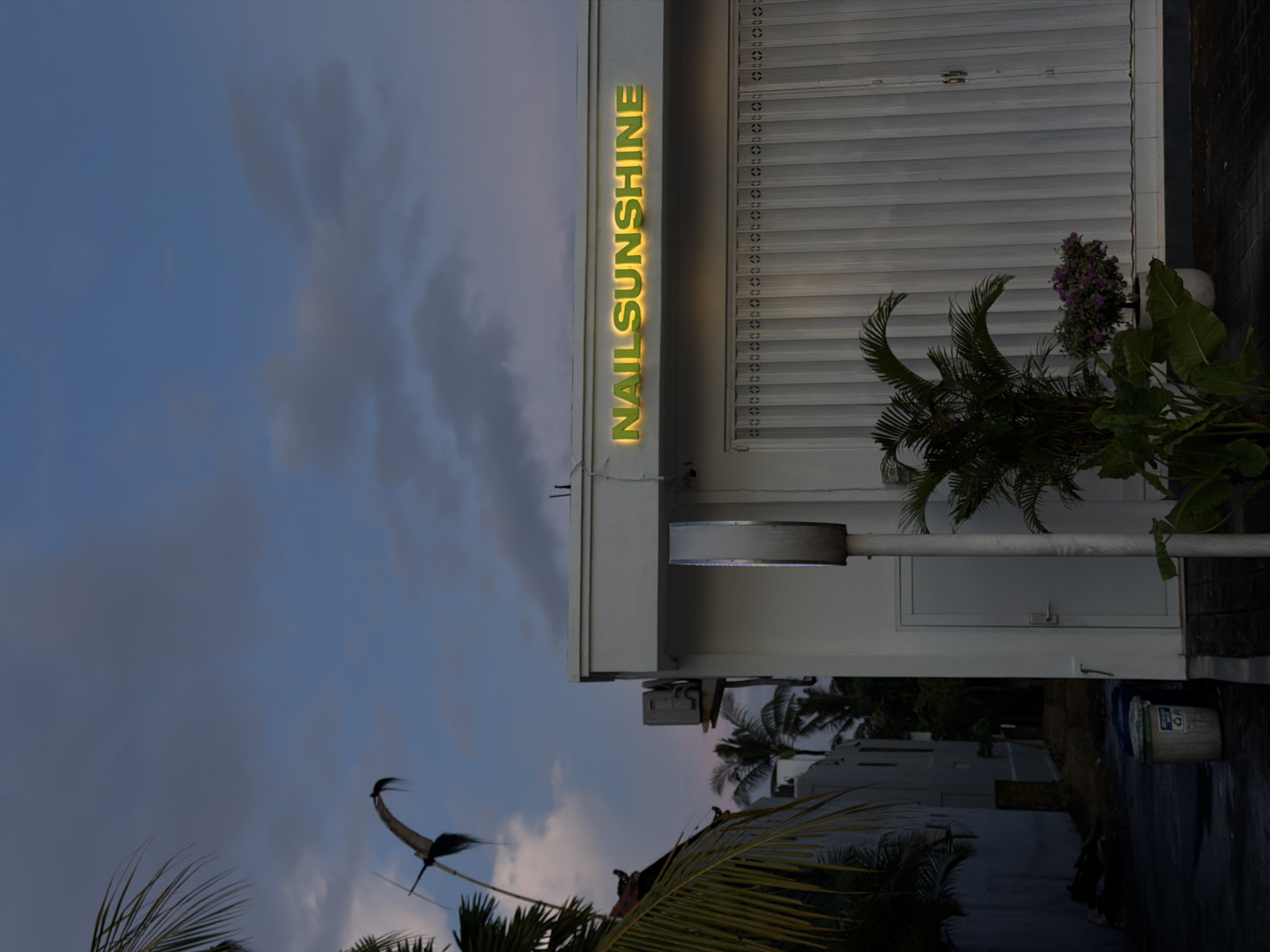 The photograph depicts the exterior of a building in Mengwi, Indonesia, during twilight. The sign "NAIL SUNSHINE" in illuminated gold lettering is affixed to the white facade. A roller shutter door and potted plants, including palm-like foliage and flowering shrubs, are situated to the right of the sign. Below the building, palm fronds are visible. A bird-like kite is visible in the sky, which is a mix of blue and grey clouds. The ground appears wet and reflects the surrounding environment. A light fixture extends from the building's facade. A bucket is visible on the ground.