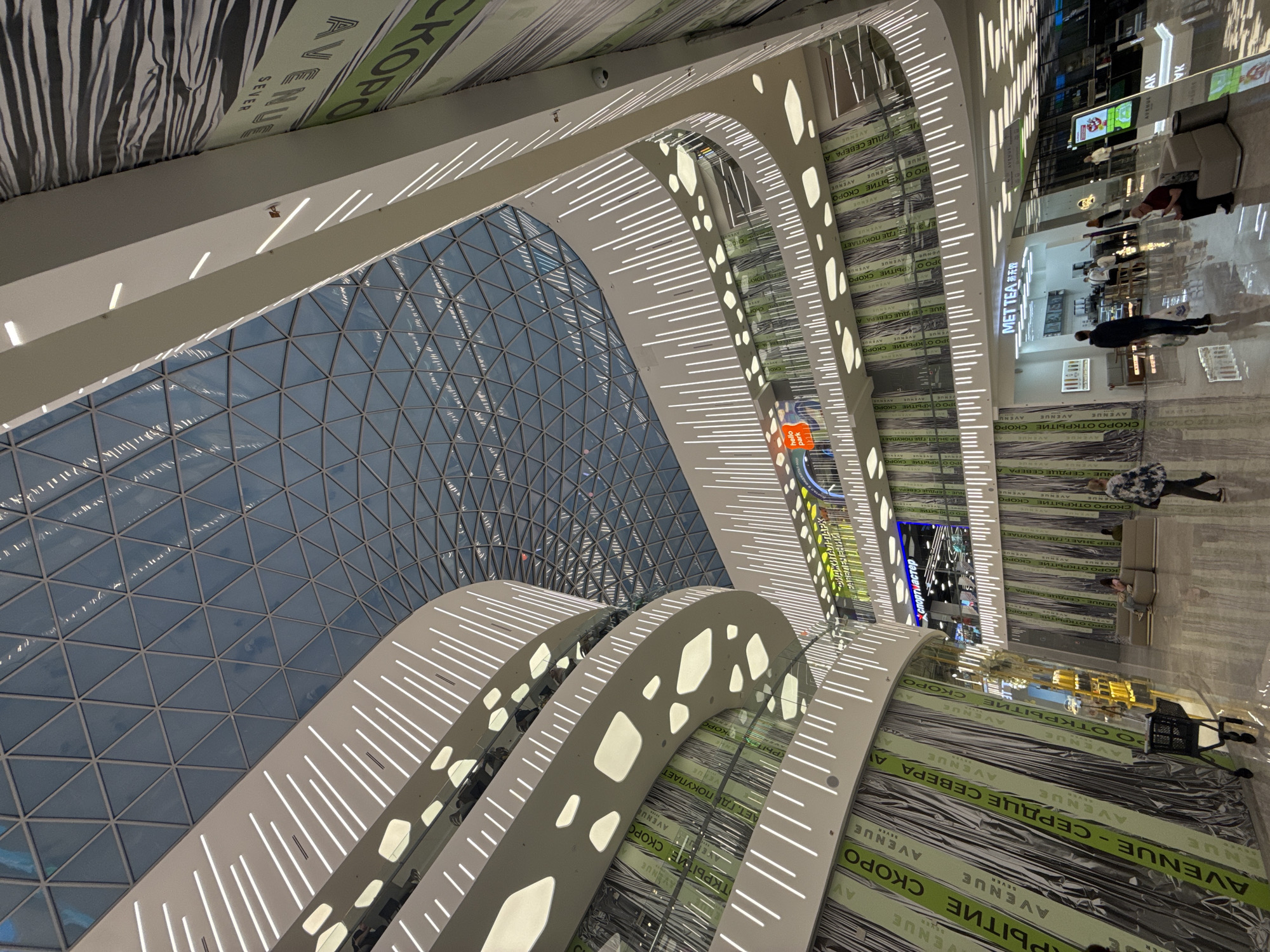 Here is a detailed description of the image:
**General Description:**
The image depicts the interior of the Aviapark shopping mall in Moscow, Russia. It is shot from an angle looking upwards, emphasizing the mall's architectural features, particularly its glass roof and layered, illuminated walkways.
**Architectural Features:**
* **Glass Roof:** The most prominent feature is a large, triangular glass roof allowing natural light to enter the space.
* **Illuminated Walkways:** The image shows layered walkways that have white lighting strips that accent the curves.
**Mall Setting:**
* **Shop Signage:** The store "Mettea" and "Спортмастер" (Sportmaster) are visible. There are advertisements that say "AVENUE - СЕРДЦЕ СЕВЕРА" (Avenue - Heart of the North).
* **Mall Furnishings:** Seating is visible.
**People:**
* **Pedestrians:** Several people are walking around the mall, and there are people lounging on the seating.