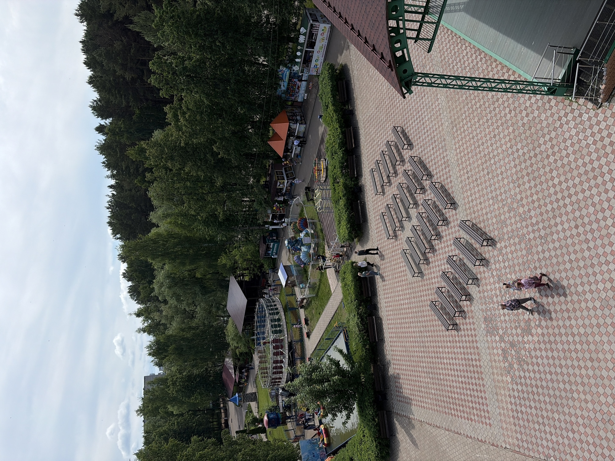 This elevated view captures an outdoor public area, likely a park or amusement park, in Chaikovsky, Russia, on an overcast daytime. The scene is dominated by a large paved plaza in the foreground, featuring a checkered pattern of light and dark reddish-brown pavers. Numerous wooden benches with metal frames are arranged in neat rows on this plaza, facing an unseen central point or stage, part of which (a green metal truss structure and a red-tiled roof) is visible in the upper right.
Three individuals are walking across the paved area in the foreground: two adults and a child. Further into the scene, a vibrant recreational area unfolds. This mid-ground section includes lush green lawns, manicured bushes lining pathways, and several amusement attractions. A prominent Ferris wheel-like ride with blue and white gondolas is visible, alongside other smaller rides and structures, some partially obscured by trees.
Interspersed among the attractions are various kiosks and stalls. One kiosk clearly advertises "МОРОЖЕНОЕ" (Ice Cream) and "НАПИТКИ" (Drinks), while another offers "СЛАДКАЯ ВАТА" (Cotton Candy), "ПОПКОРН" (Popcorn), "НАПИТКИ" (Drinks), and "ЧУПА-ЧУПСЫ" (Chupa Chups), indicating typical park refreshments. A small body of water, possibly a pond or canal, features a yellow inflatable boat. A fountain is also visible near one of the kiosks.
The background is defined by a dense line of tall green trees, suggesting a forest or extensive wooded area bordering the park. The sky is largely overcast with some visible clouds, and the shadows cast by the benches indicate it's daytime, likely late morning or afternoon. The overall atmosphere appears calm and leisurely.