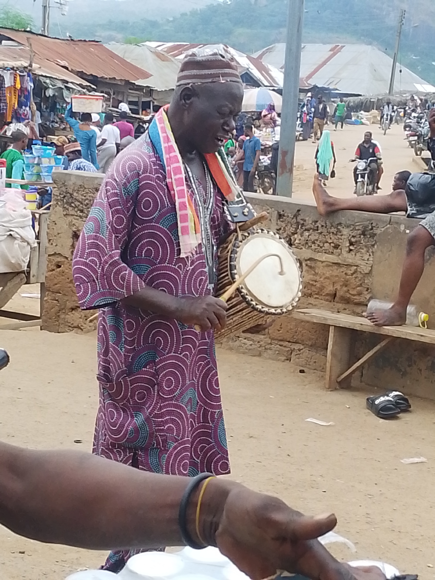 A man in a traditional purple and blue patterned tunic and a striped hat plays a drum in what appears to be an outdoor market in Komu, Nigeria. The man holds a drumstick in his right hand, poised to strike the drum. The drum is cylindrical with a stretched white skin on one end and is decorated with horizontal bands of woven material. He wears a striped scarf around his neck. In the background, stalls with corrugated metal roofs are visible, lined with goods. Various people are present, some walking and others sitting. A motorbike is parked on the right side of the frame. The ground is dirt. A person's arm is in the foreground, with a black and yellow bracelet on the wrist.