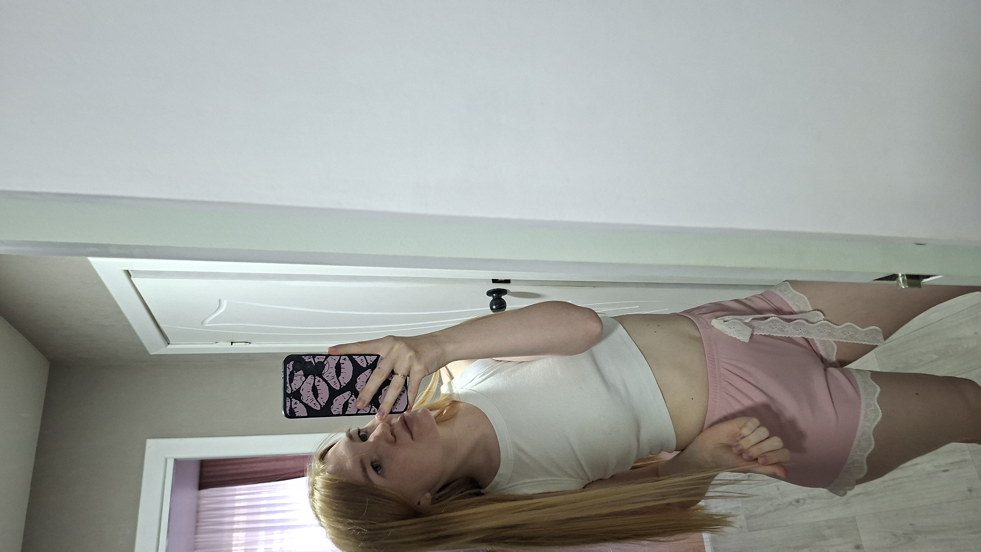In a bedroom, a young woman with long blonde hair, wearing a white crop top and pink pajama shorts, takes a selfie in a full-length mirror. The room has light gray walls and a white door. A curtained window with pink drapes is visible in the background. The scene is likely set in Chaikovsky, Russia.