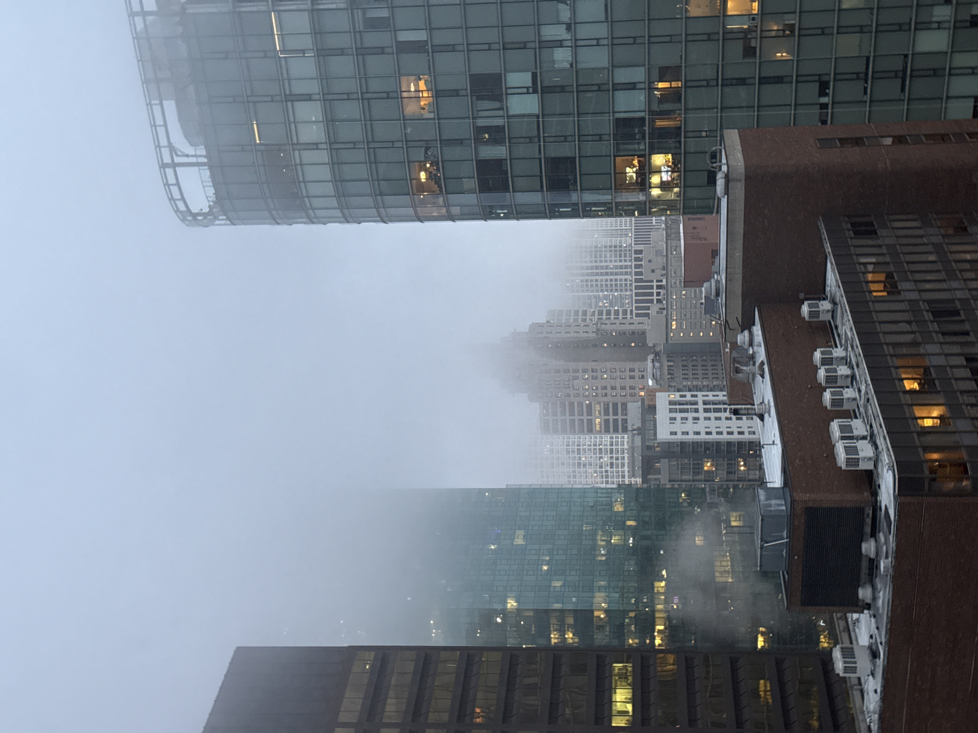 This is an aerial, nighttime, outdoor shot of a dense urban cityscape in Chicago, USA. The image is dominated by modern skyscrapers, many with glass facades that reflect the city lights. The scene is partially obscured by a thick layer of fog or mist, creating a hazy and somewhat ethereal atmosphere.
The foreground features a dark, brick-clad building with several exposed air conditioning units visible on its roof. To the left and slightly behind this, a sleek, curved glass skyscraper rises, its grid-like structure illuminated from within. Further into the background, a multitude of other buildings are visible, their lights twinkling through the fog, creating a sense of depth and the vastness of the city.
The time of day is clearly night, indicated by the artificial lights emanating from the buildings. The weather is foggy, which significantly impacts the visibility of the cityscape and contributes to the overall mood of the image. There are no visible people in the frame, but the lit windows suggest occupied spaces within the buildings. The overall emotion conveyed is one of urban grandeur and mystery, amplified by the obscuring fog. No text is visible in the image.