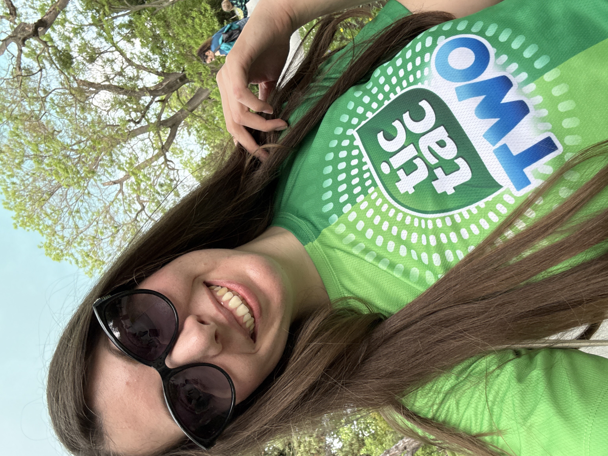A young woman with long brown hair and sunglasses smiles at the camera while wearing a green t-shirt with a "Tic Tac Two" logo. She is outside, likely in a park, with trees and a blue sky in the background. Other people can be seen indistinctly in the background. The location is Unknown, Unknown.