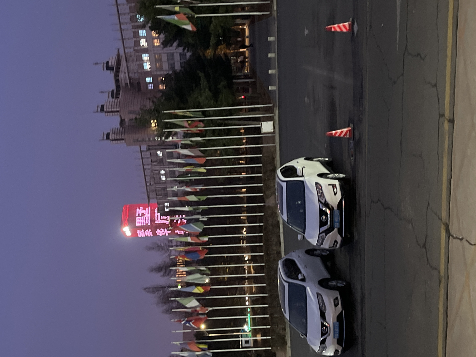The image depicts a street scene in Changchun City, China, at dusk. On the left, a tall building is visible, along with a large red LED sign displaying Chinese characters. Rows of flagpoles with various national flags line the side of the road. Two white cars are parked on the right side of the street, parallel to the curb. Two orange and white striped traffic cones are positioned on the road further back. The sky is a dark shade of blue, indicating early evening. The street is paved and shows signs of wear. There are no people visible in the image.
