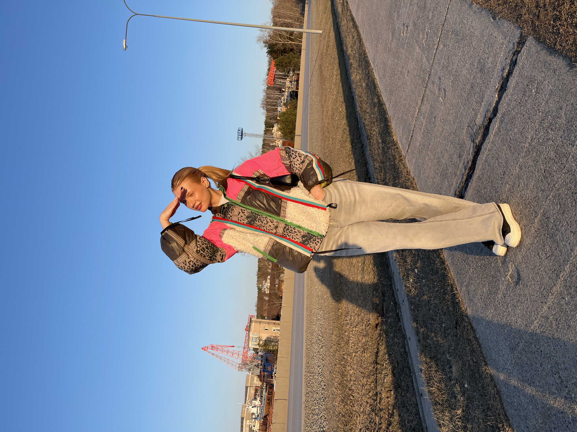 The photograph depicts a young woman standing on a sidewalk in what appears to be Chaikovsky, Russia. She is shielding her eyes with her hand, possibly from the sun. She is wearing a patterned jacket, light-colored jeans, and sneakers. Behind her, across the road, is a waterfront area, including an orange crane and industrial buildings, indicating a port or shipyard. The sky is clear and blue. A street lamp is in the upper left corner of the frame. The sidewalk edge is lined with dry grass.