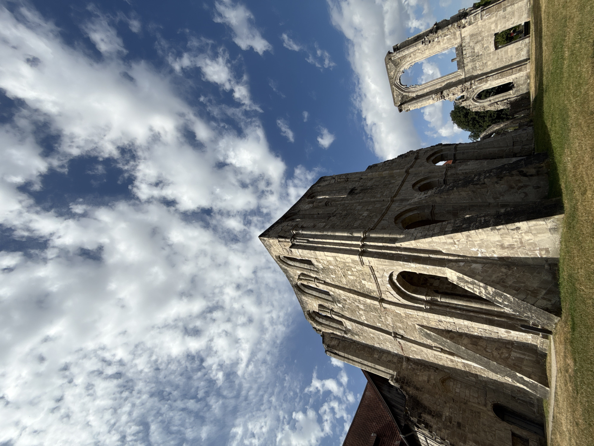 The image shows the ruins of the Walkenried Abbey in Walkenried, Germany, on a sunny day with a bright blue sky dotted with white clouds. The foreground is a grassy area with dry patches of straw. Two main structures of the abbey ruins are visible. On the left, a large, imposing stone building stands with arched windows and buttresses. A portion of a red-tiled roof is visible to the left of this structure. To the right, a second, more fragmented ruin is present, featuring a large, Gothic-style arched opening. Trees are visible behind the ruins in the distance. There are no people or animals in the image. The overall impression is one of historical grandeur and decay.