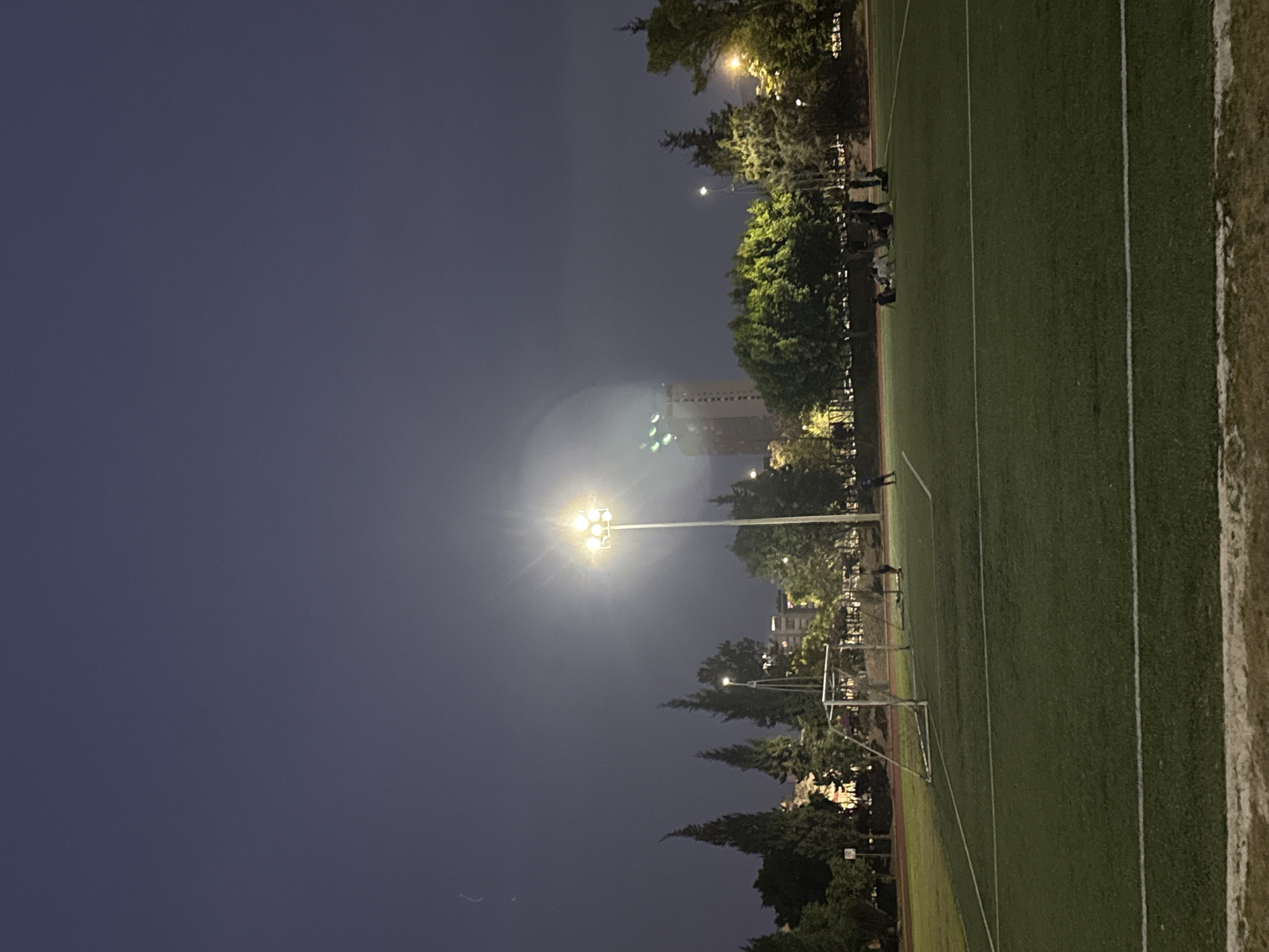 The photograph shows a soccer field at night. The field is green with white lines marking the boundaries. A tall light post illuminates the field, and a soccer goal is visible at one end. Trees surround the field. In the background, a building is visible. A few people are standing on the edge of the field. It is impossible to know the precise location in Puebla, Mexico from this photograph.