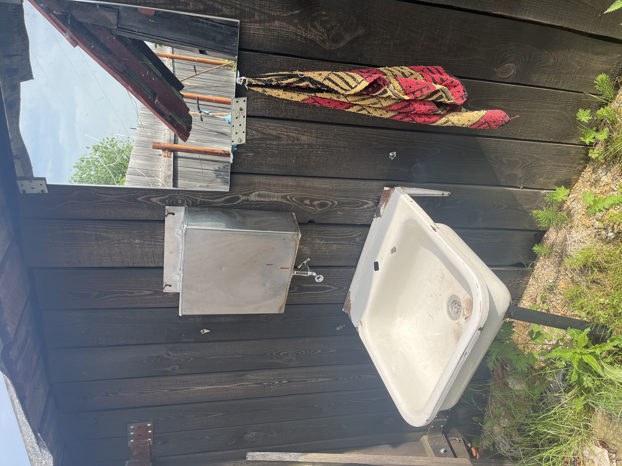 The image depicts an outdoor sink and a mirror mounted on a dark wooden wall. The sink is white, porcelain, and appears aged, with rust spots around the edges and faucet. A metal water tank is mounted above the sink, with a silver faucet attached. A towel with a red, yellow, and black striped pattern hangs from a hook to the right of the mirror. The mirror itself is rectangular and reflects an outdoor scene with a wooden fence, trees, and a cloudy sky. The area around the sink is overgrown with grass and weeds, suggesting it's in a rural or neglected outdoor setting. Based on the provided information, this scene is located in the city of Berezovskii, Russia.