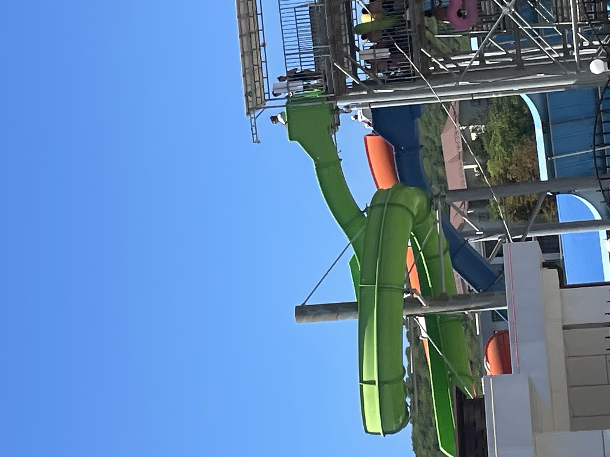 In Dzhubga, Russia, a bright blue sky serves as the backdrop for a water park. The main focus is a multi-level structure with various water slides. A prominent, large, bright green slide dominates the left side of the frame, curving downward. Below it, an orange slide is visible, and further down, a blue slide.
People are visible on the upper levels of the water park structure, some standing near the entrance of a slide and others on a walkway. One individual in a white shirt is at the top of the green slide. Several other people are seen on a platform with railings, and some are on stairs. On the far right, a pink inflatable ring is attached to the structure.
The environment includes lush green foliage and buildings in the background, suggesting a hillside setting. The water park structures are made of metal and plastic. The overall scene depicts a sunny day at a recreational facility.