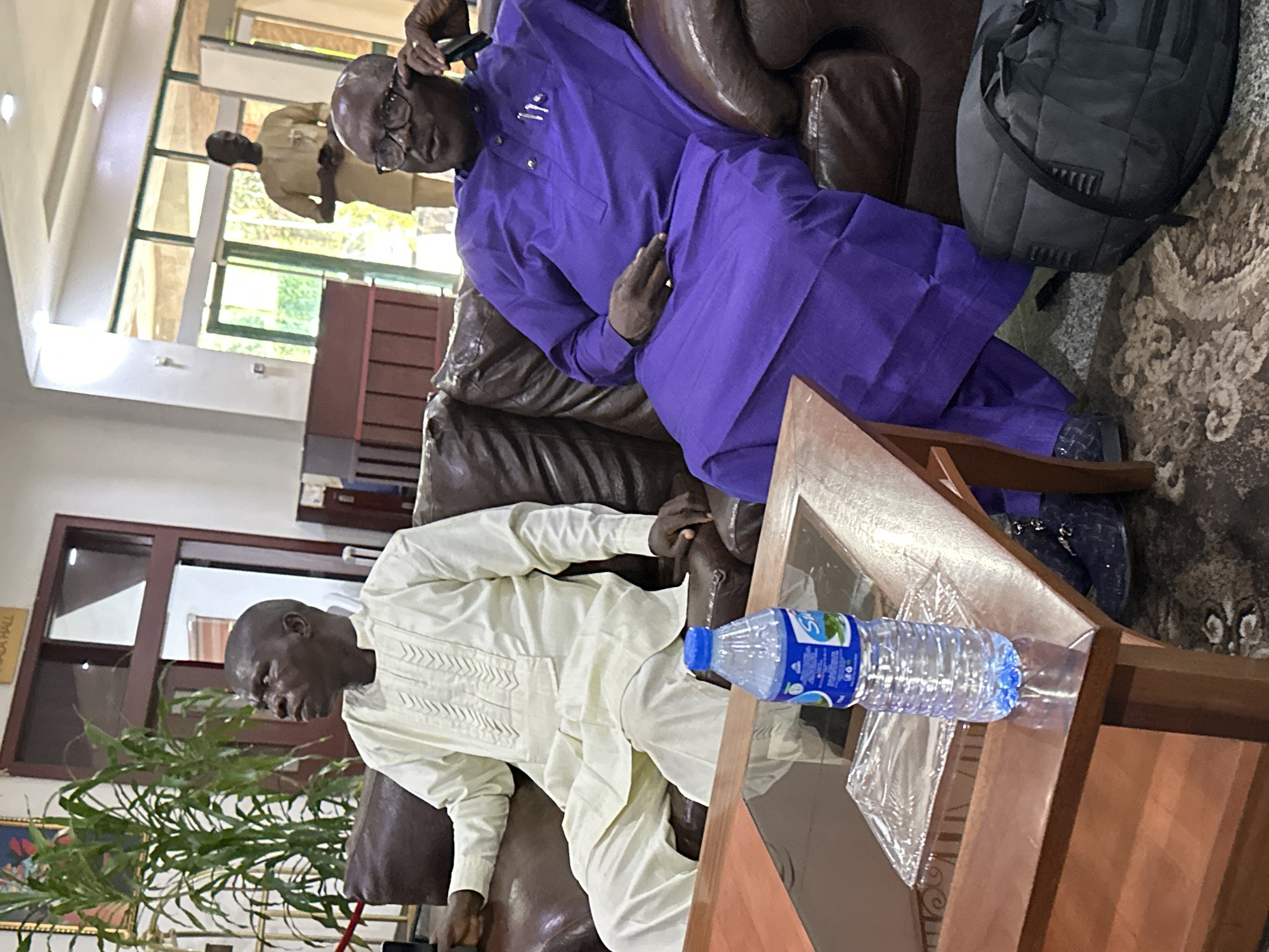 In Abuja, Nigeria, two men are seated on a brown leather sofa in what appears to be a waiting area or lounge. The man on the left wears a cream-colored traditional outfit, looking upwards and to the left. The man on the right is dressed in a vibrant purple traditional outfit, wearing glasses and resting his chin on his hand, looking towards the right. A wooden coffee table is positioned in front of the sofa, holding a bottle of water and a clear plastic container. In the background, a third man in a light brown outfit stands near large glass doors, and a tall potted plant is visible to the far left. The overall setting suggests an indoor, communal space within a building.
