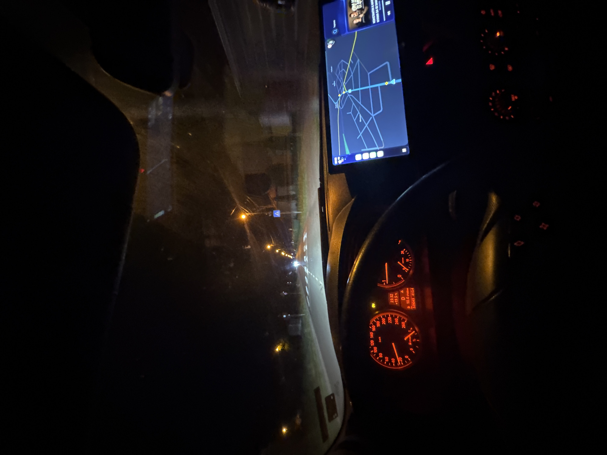 The image depicts the interior of a car at night, likely in transit. The dashboard is visible, with illuminated gauges showing a speed of approximately 15 km/h, and the time is 23:32. A navigation screen displays a map, possibly guiding the driver. The environment appears to be a road with streetlights. The OCR text suggests the car is in the vicinity of "KONSTANTIN & MAR," and a distance of "10.4 1200km" is displayed. No people are visible. Given the information, it's plausible the car is in or near Mogila, Bulgaria, based on the identified location on the navigation screen.