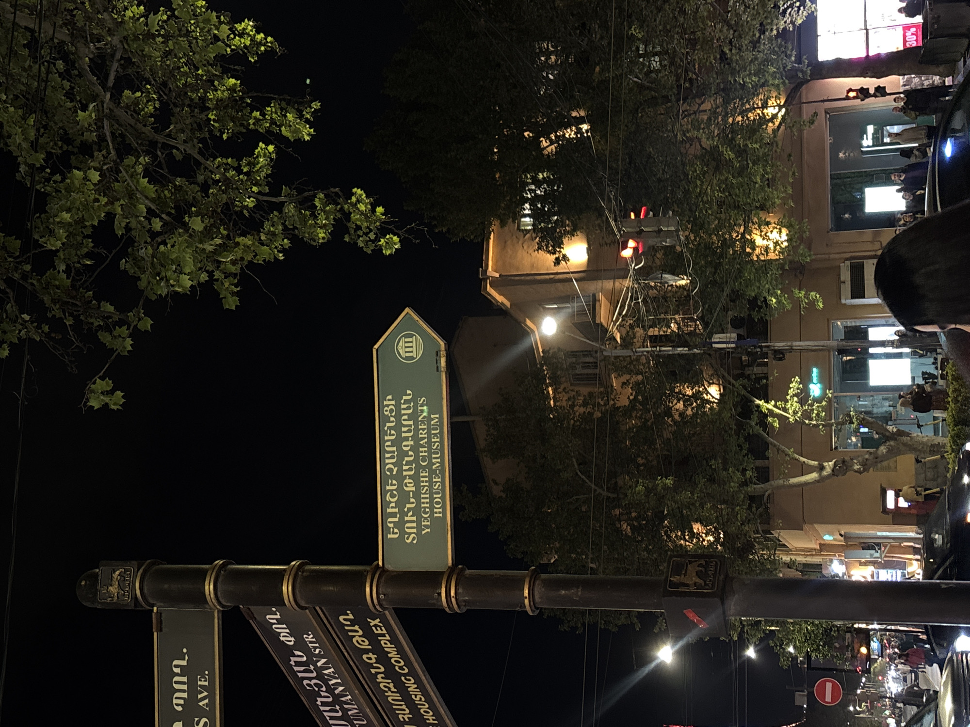 The image shows a street scene at night in Yerevan, Armenia. In the foreground is a street sign with directions in both Armenian and English. The sign points to the Yeghishe Charents House-Museum, and also indicates directions for Tumanyan Str. and Housing Complex. The sign is mounted on a dark metal pole with decorative elements.
Behind the sign, a street with cars and pedestrians is visible. The buildings along the street are illuminated, with light spilling onto the sidewalk. Trees with green leaves partially obscure the view of the buildings.
In the background, storefronts with bright lighting illuminate the night sky. The scene suggests a lively urban environment.