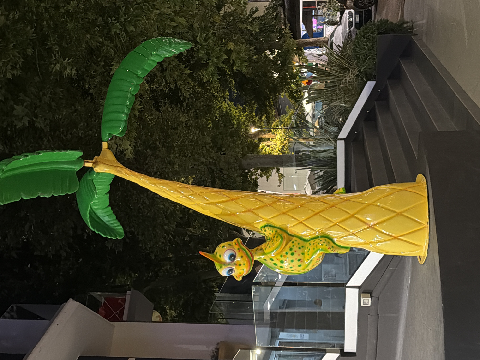 The image shows a brightly colored statue of a dinosaur climbing a yellow, palm-like structure with green leaves at the top, located in Gatteo, Italy. The dinosaur is predominantly green with yellow and red spots and has large, cartoonish eyes. The palm structure is textured with a diamond pattern and sits on a dark gray set of stairs. Behind the statue, there are trees with lush green foliage, some of which are illuminated by streetlights. A building with glass windows and a white railing is visible to the left. In the background, cars and outdoor seating areas suggest a commercial or entertainment district. The overall scene is well-lit, indicating it is nighttime or dusk, with artificial lighting illuminating the statue and surrounding area.