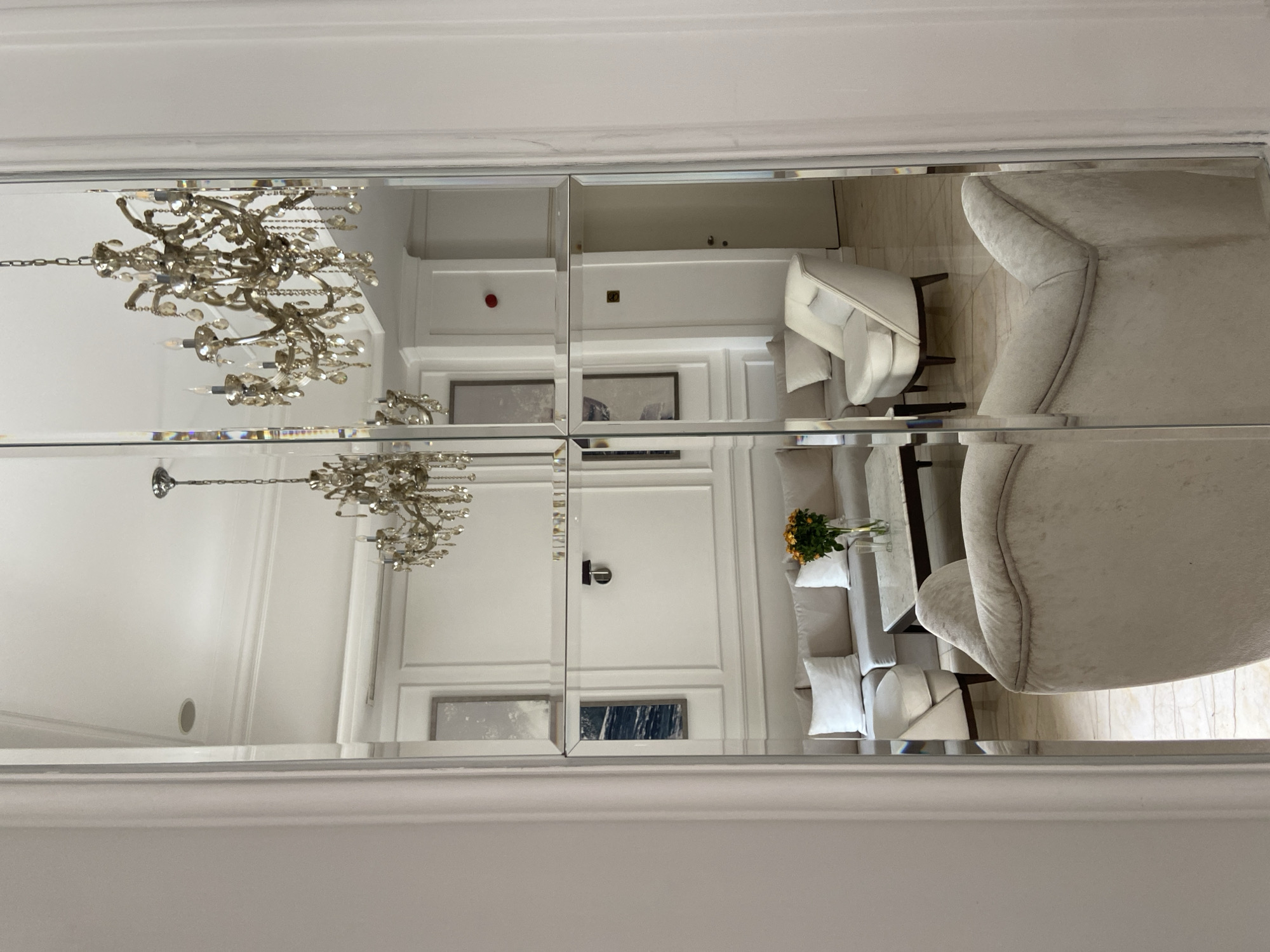 This image captures a luxurious and elegantly appointed indoor space, likely a hotel lobby or a refined living area, viewed through a large, segmented mirrored wall. The overall aesthetic is bright and clean, dominated by white and light neutral tones.
The central feature visible in the reflection is a grand, multi-tiered crystal chandelier, showcasing intricate metallic work and numerous sparkling crystals, hanging from a high white ceiling adorned with molding.
The mirrored surface reflects various elements of the room, including plush, light-colored upholstered armchairs, one of which has distinct winged sides, and a sofa or bench adorned with white throw pillows. A coffee table, possibly made of light marble or polished stone, holds a vase containing a vibrant bouquet of yellow flowers, providing a subtle splash of color.
In the background of the reflection, white paneled walls are visible, featuring framed artwork and subtle architectural details. A small red fire alarm button and a discreet gold plaque are also reflected on one of the walls. Several white doors with dark, sleek handles contribute to the contemporary yet classic design.
No people are present, and no specific activity is taking place, lending the scene a serene and undisturbed atmosphere. The bright illumination suggests it is daytime, or the space is well-lit by interior lighting. The sophisticated decor, high-end furnishings, and pristine condition evoke a sense of luxury and tranquility, fitting for a premium location like Budva, Montenegro. There is no visible text in the image.