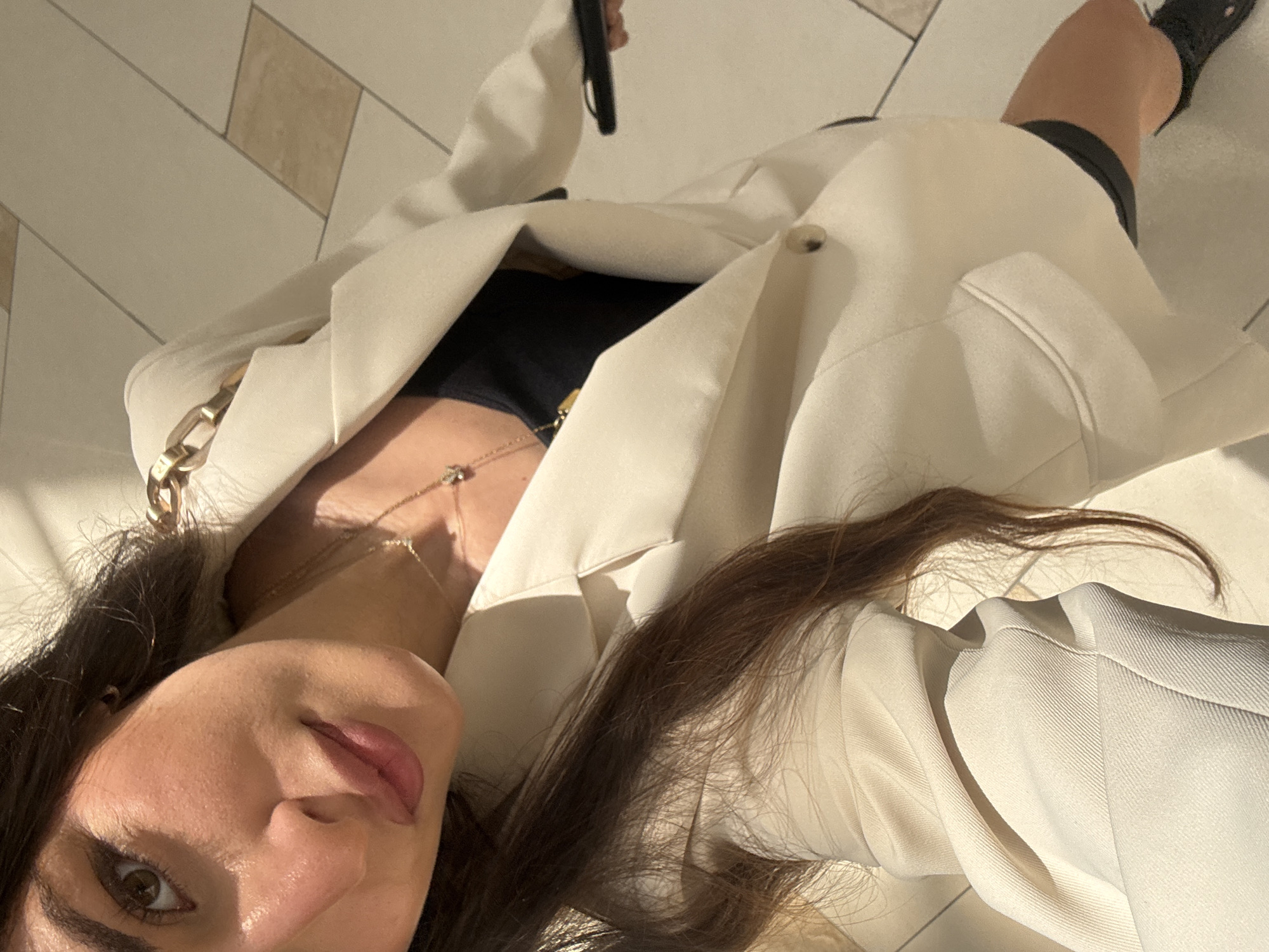 A close-up, high-angle shot depicts a woman lying on a light-colored tiled floor in what appears to be an indoor location, possibly a shopping mall in Aventura, Florida, USA. She is wearing a large, off-white blazer over a black top, with a gold chain accenting her blazer. A delicate gold necklace is visible around her neck. She is also wearing black shorts and black shoes. She is holding a black mobile phone in one hand. The lighting is bright and slightly diffused, casting shadows and highlights across the scene.