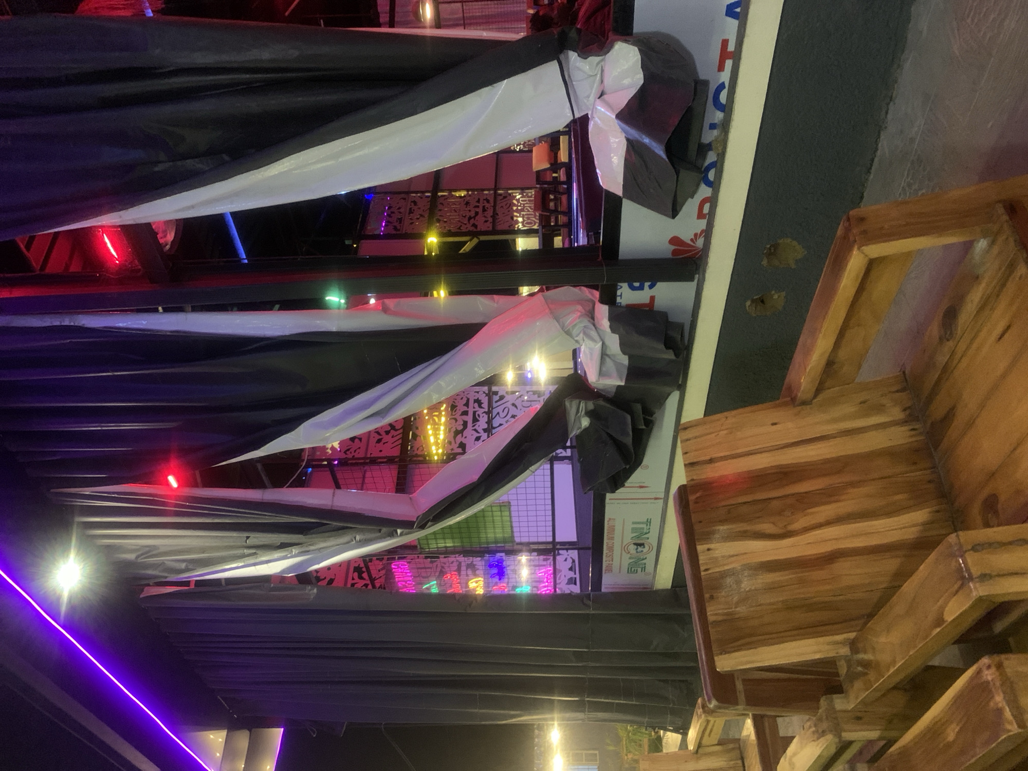 The image depicts an outdoor seating area at night, likely a restaurant or bar, on Rukuba Road, Nigeria. The foreground features rustic wooden chairs and benches arranged in a multi-level seating configuration. Behind the seating, dramatic black and white curtains are draped, creating a visually striking backdrop. Beyond the curtains, the scene opens up to reveal neon lights in various colors, including pink, green, blue, and yellow, illuminating decorative panels and suggesting a vibrant nightlife atmosphere. The lighting is primarily artificial, with a prominent purple neon strip running along the top edge of the frame and red lights casting glows from within the interior. The ground appears to be paved, with some areas showing a grid-like pattern. The overall impression is one of a contemporary urban entertainment venue.