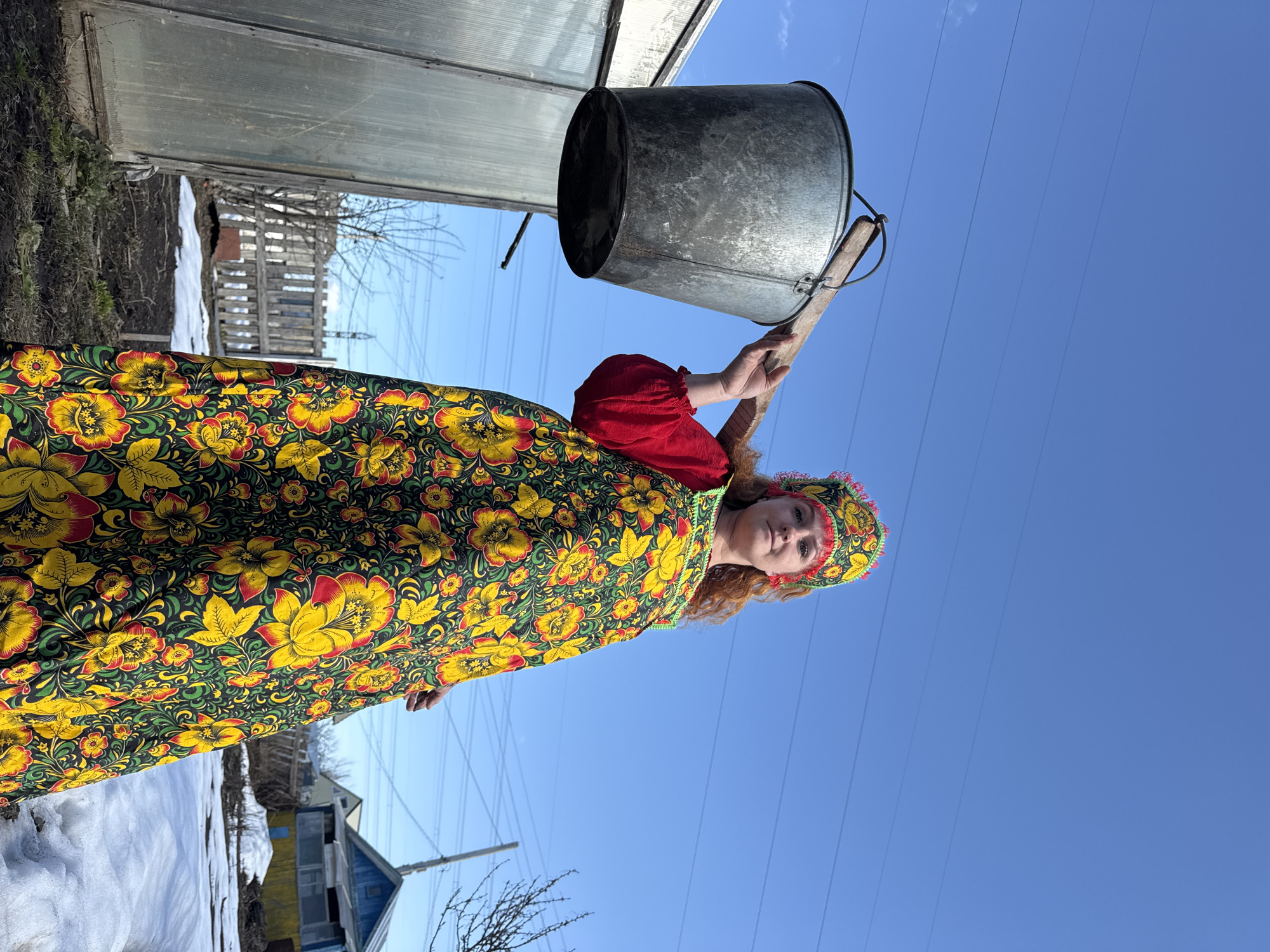A woman dressed in traditional Russian clothing stands outside with a bucket, possibly in Chaikovsky, Russia. The woman is wearing a colorful floral dress, a red blouse, and a matching headpiece. She is holding a metal bucket on a wooden yoke. The background includes a blue sky, utility lines, patches of snow, and buildings. There is a greenhouse or similar structure adjacent to the woman.