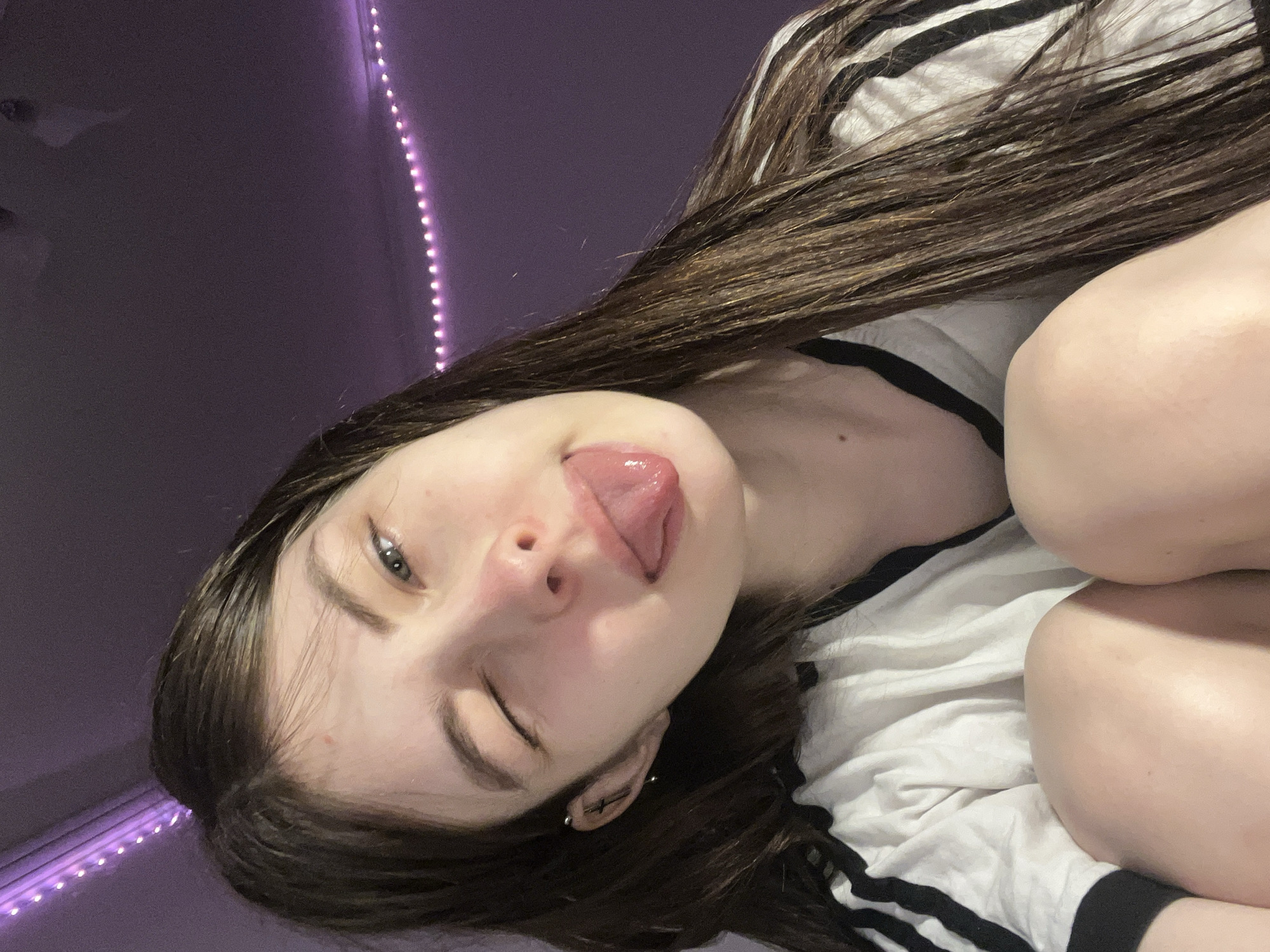 A young woman is photographed indoors. She is positioned at an angle, facing the camera, with her head tilted to the left. She has long, straight, brown hair and is wearing a white and black striped shirt. Her left eye is closed in a wink, and her tongue is extended slightly out of her mouth. She has a fair complexion and appears to have ear piercings. The background consists of a purple wall with a string of LED lights running vertically along the edge of the frame. There is no evidence to indicate this photo was taken in Saint Petersburg, Russia.