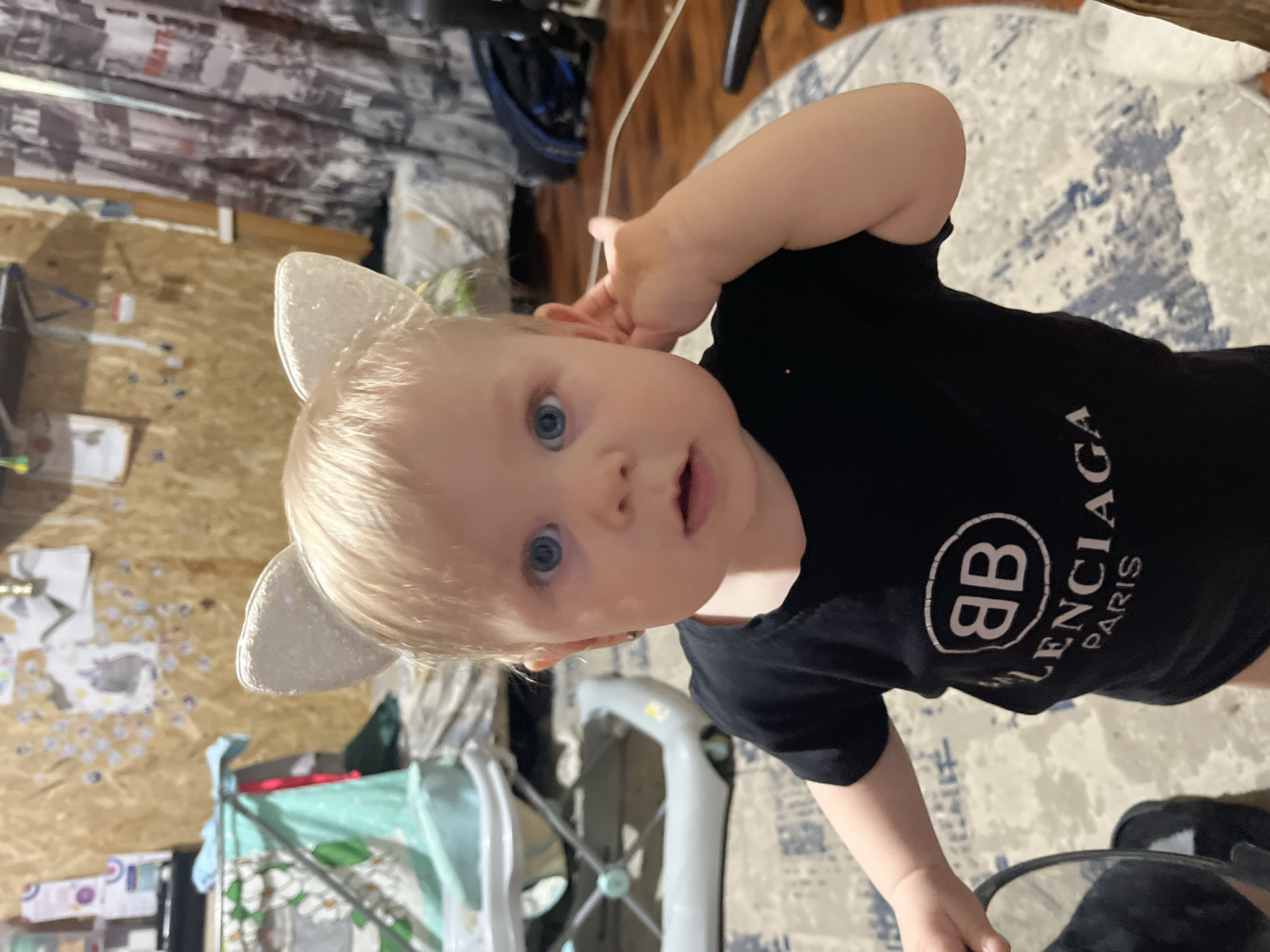 A baby with blonde hair and blue eyes is wearing silver cat ears and a black t-shirt with the "Balenciaga Paris" logo. The baby is pointing to their ear with their left hand. The background includes a carpet with a blue and white pattern, a baby walker, and a wall with drawings. There is also a chair and some miscellaneous items in the background. The location is likely inside a home and there is no indication of Vesyoloye, Russia.