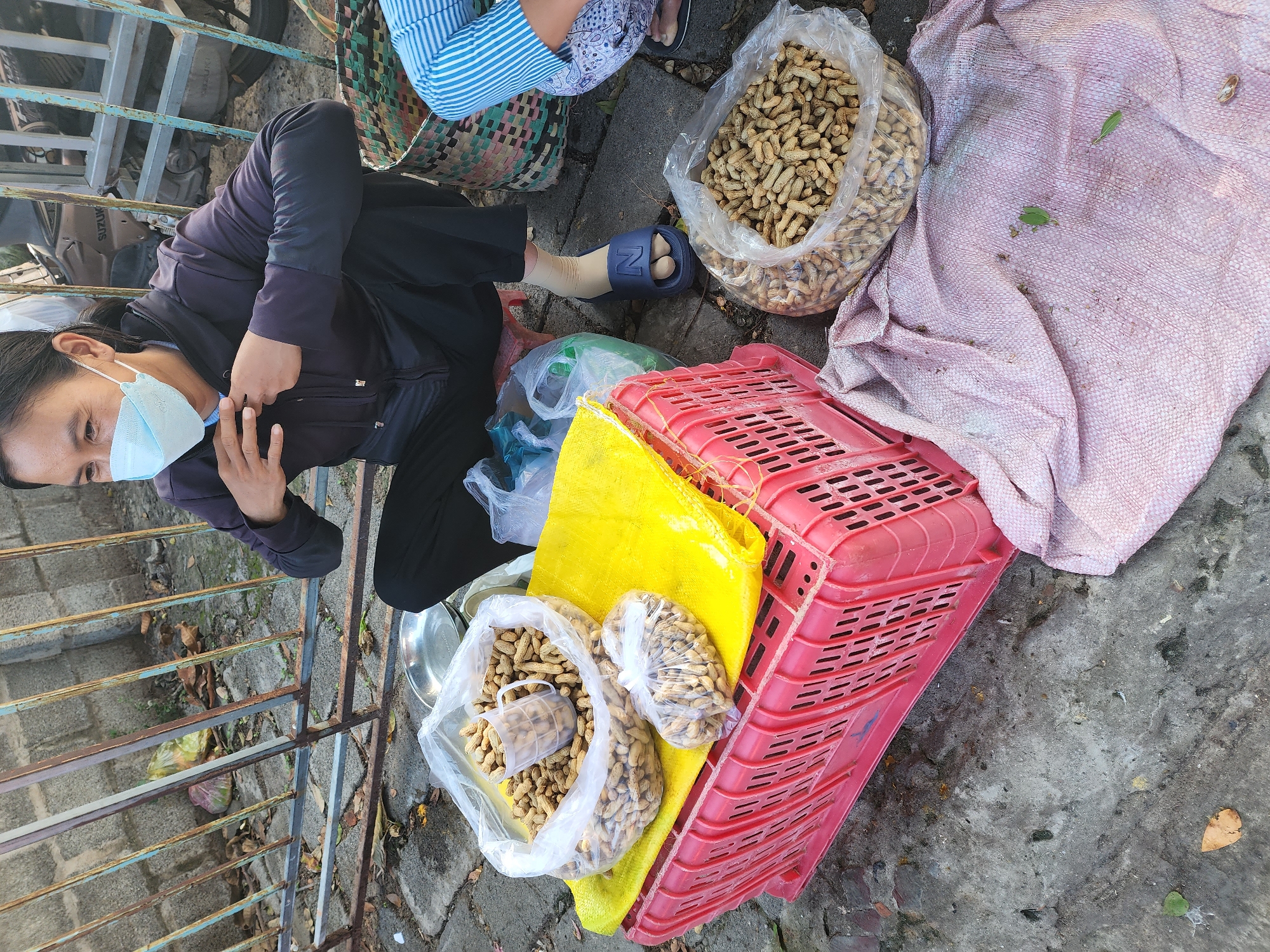 In Phu Thuy Ward, Vietnam, a woman wearing a blue face mask and a dark jacket sits on a stool on a paved area, possibly a street or market. She is reaching towards her chest. In front of her, a large red plastic crate holds a yellow cloth, upon which sit two plastic bags filled with peanuts. One bag is larger, with a clear plastic cup inside, and the other is smaller. To the right of the crate, another large plastic bag of peanuts rests on a pinkish cloth spread on the ground. A woven basket is visible to the right of the woman, and a person wearing striped pants is partially visible in the lower right corner. A metal fence with rusty bars is behind the woman, and a motorcycle is partially visible through the fence. The ground is made of uneven paving stones and concrete.