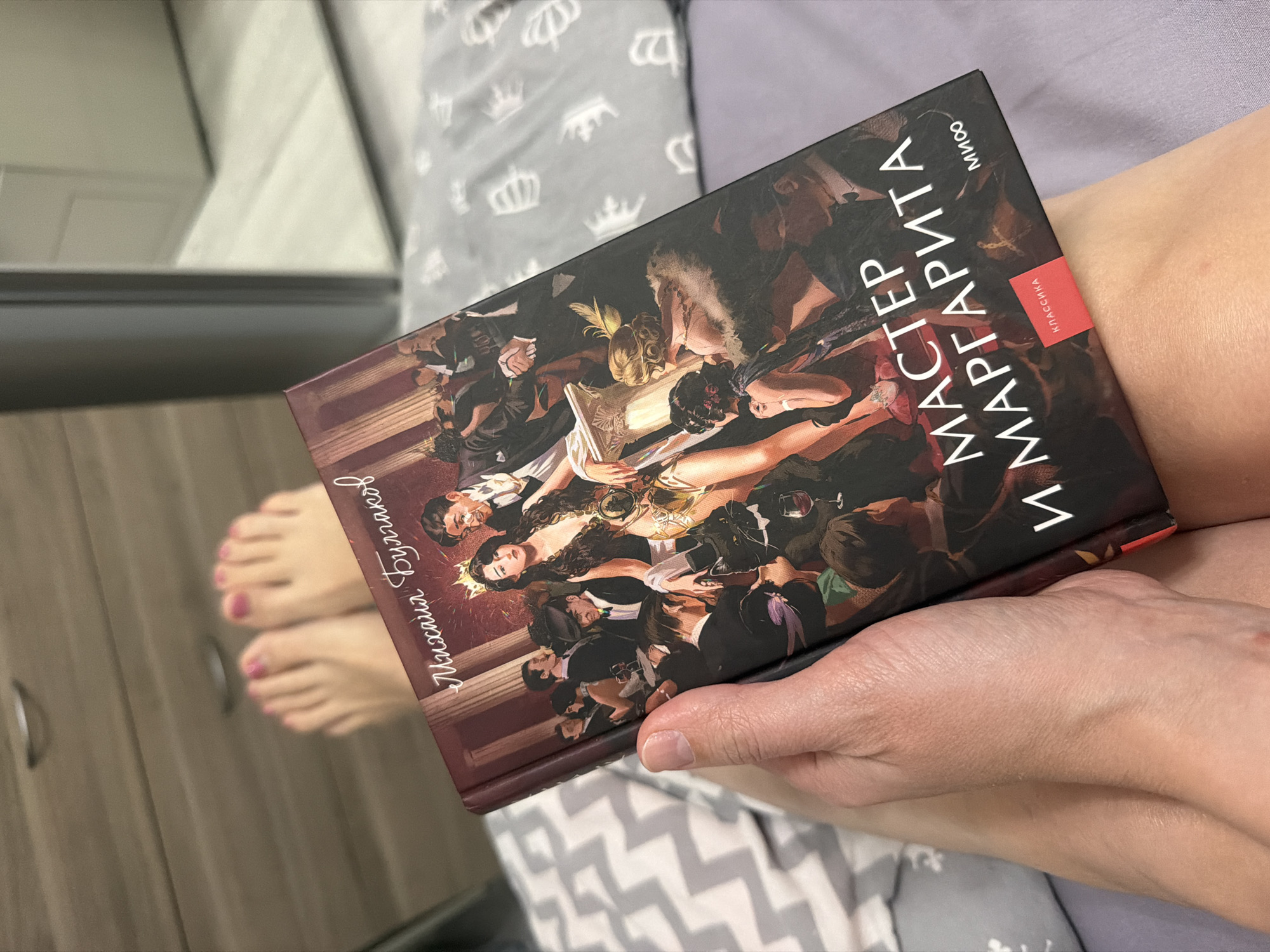 An indoor scene in Rostov-on-Don, Russia, depicts a person reclining in bed, holding an open copy of Mikhail Bulgakov's novel, "Master and Margarita." The book's cover features a detailed illustration of a lavish, chaotic party, including a central female figure wearing a crown and gold attire, a man in a tuxedo, and a black cat in a bowtie holding a wine glass amidst other formally dressed individuals and classical columns. The book's spine is reddish-brown, and the cover includes a red "CLASSICS" label and "MIF" publisher logo. The person's right hand grips the book from the underside, revealing natural fingernails. In the blurred background, the person's bare feet are visible, adorned with pink nail polish, resting against a light-colored wooden dresser with two silver handles. The bed is covered with two distinct patterns of bedding: a grey and white zigzag pattern on the left and a solid light purple fabric on the right, beneath the person's upper legs. A blurry reflective surface or mirror is visible in the upper right background.
