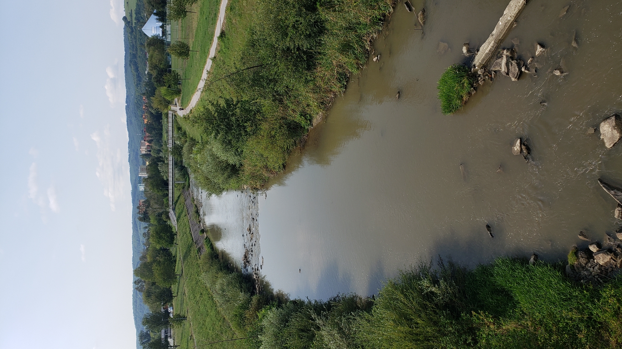 This is an aerial view of the Torysa River flowing through the city of Prešov, Slovakia. The river is murky brown with some debris and rocks visible on the surface and along its banks. Lush green vegetation lines both sides of the river, with trees and bushes growing densely. A paved pathway runs along the right bank, and a pedestrian bridge spans the river in the distance. In the background, there are residential buildings and a hilly landscape with more trees. The sky is partly cloudy, and the overall scene suggests a sunny day.