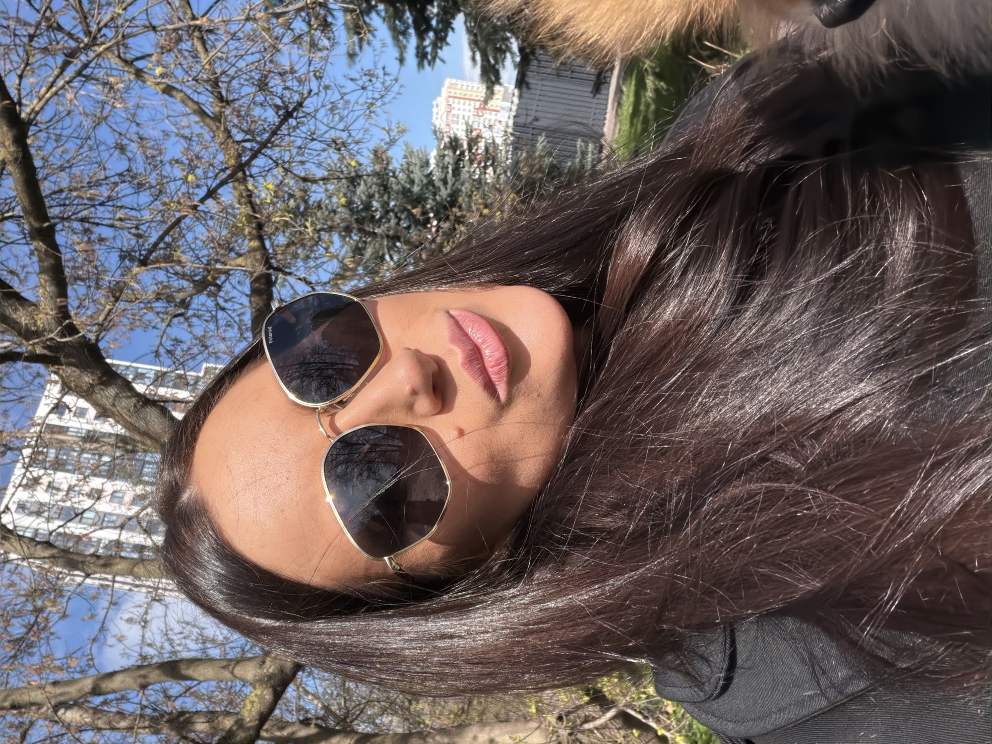 A young woman poses for a selfie outdoors, tilted slightly to the left. She is wearing sunglasses with gold frames that reflect the sky and trees. Her long, dark hair is glossy and falls over her shoulder. She has light skin and pink lipstick. In the background, bare tree branches reach towards a clear blue sky. A building with white facades is visible behind the trees. Given the prompt, the location is likely in Khimki, Russia.