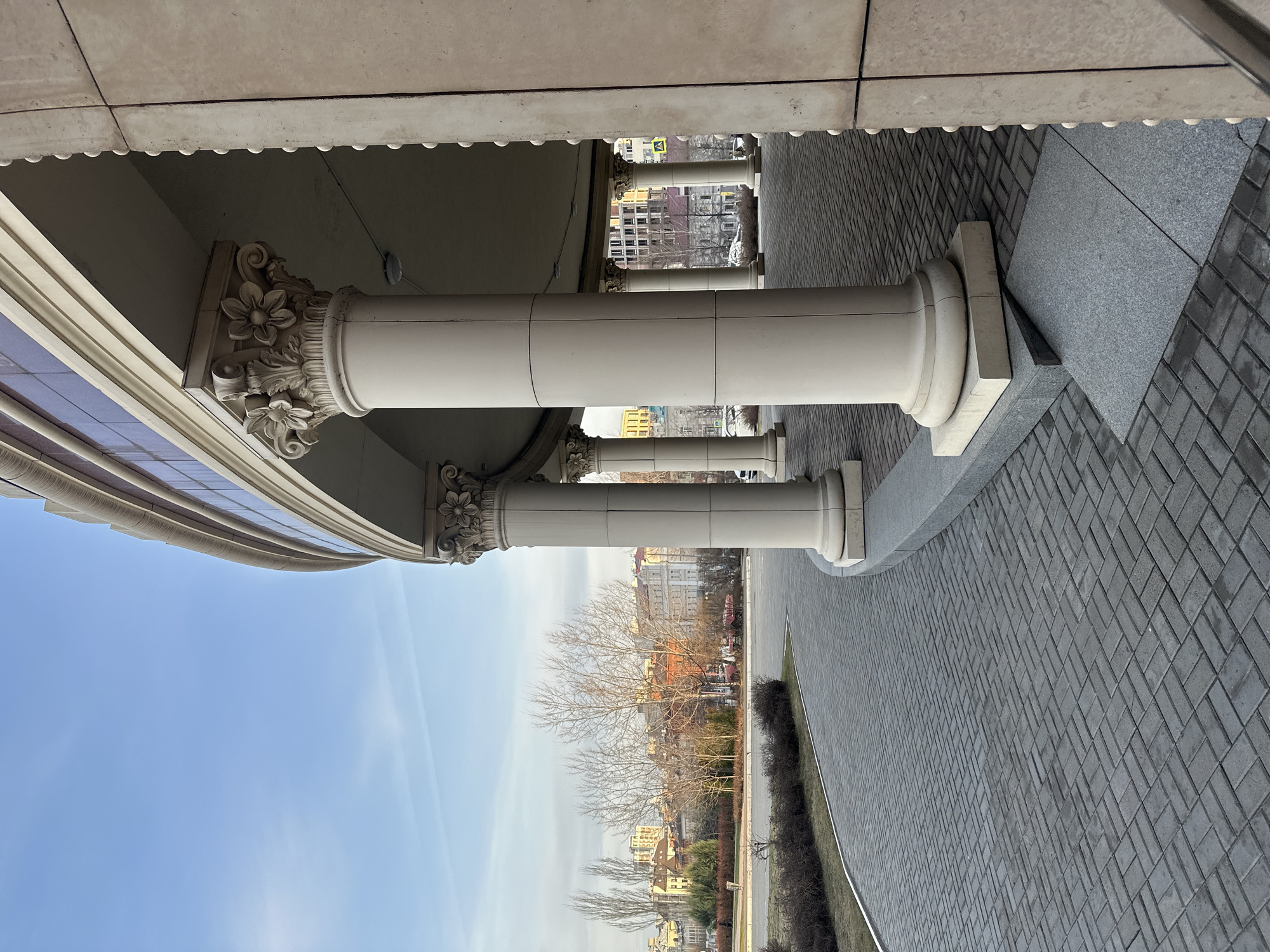 The image shows a row of white, cylindrical columns with ornate, floral capitals supporting an overhanging structure. The columns are spaced evenly along a paved walkway made of gray rectangular blocks. Beyond the columns and walkway is a view of the city of Kazan, Russia, with buildings and bare trees visible under a blue sky with wispy clouds. A thin line of vegetation borders the walkway. The perspective is slightly angled, looking upwards towards the structure and columns.