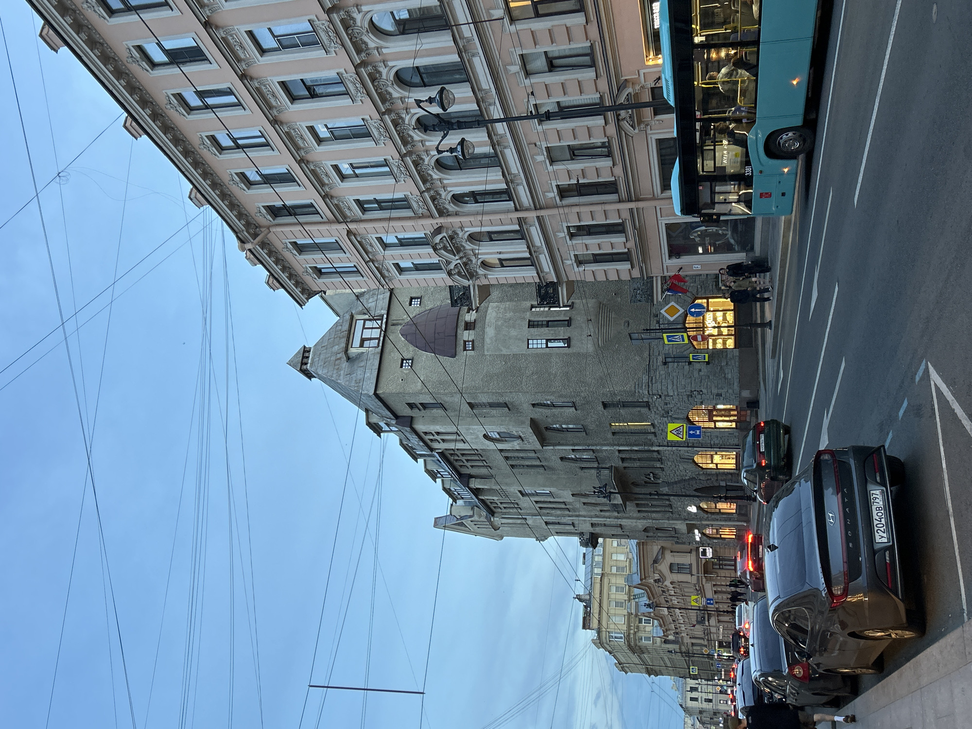 The image depicts a street scene in a city during dusk or early evening. The sky is a soft blue, with numerous overhead wires crisscrossing the frame. On the right side of the street, a tall, light pink building with ornate architectural details and numerous windows stands prominently. To its left, a taller, grey building with a distinctive pointed roof and many windows dominates the mid-ground. Further down the street, more buildings with various architectural styles are visible.
The street itself is paved and marked with white lane lines. Several cars are parked along the sidewalk on the left side of the image, and a line of cars is driving on the road. A bright blue bus is visible in the right lane, with passengers visible inside. People are walking on the sidewalk and crossing the street in the distance. Yellow traffic signs with black symbols are posted along the road. The overall atmosphere is that of a bustling urban environment transitioning into the evening.