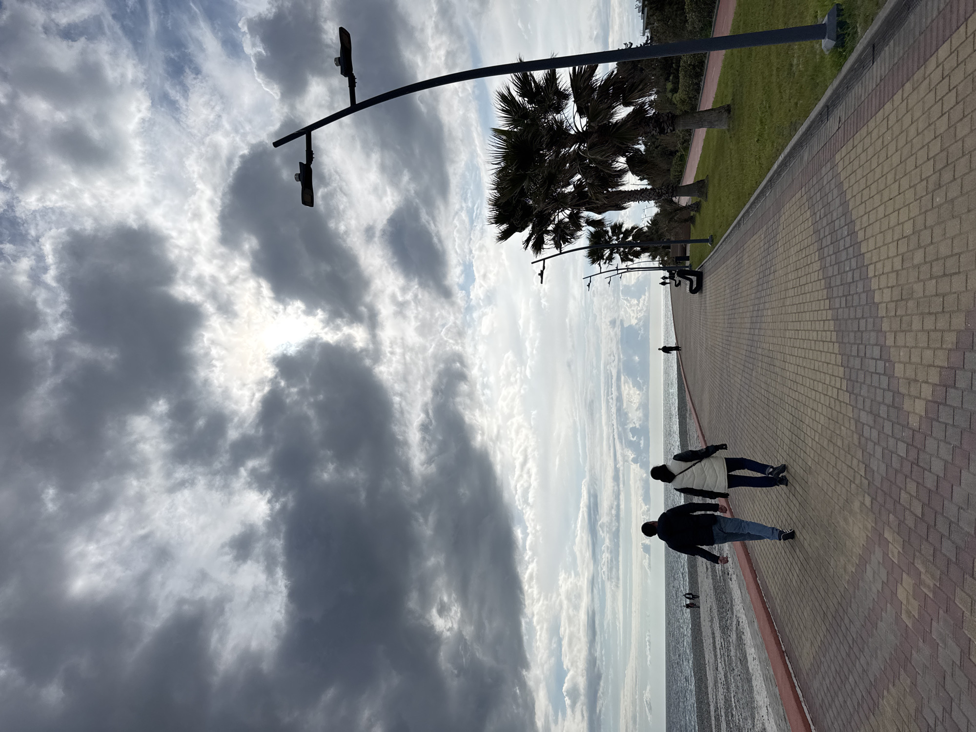 The image depicts a seaside promenade in Sirius, Russia. The sky is filled with a mix of dark and light clouds, partially obscuring the sun. Along the brick-paved walkway, there are palm trees and lampposts. Two individuals are walking, with one wearing a dark jacket and jeans, and the other a white jacket. A single person stands in the distance. The promenade is adjacent to a green strip of grass, possibly a park. The water extends into the horizon.