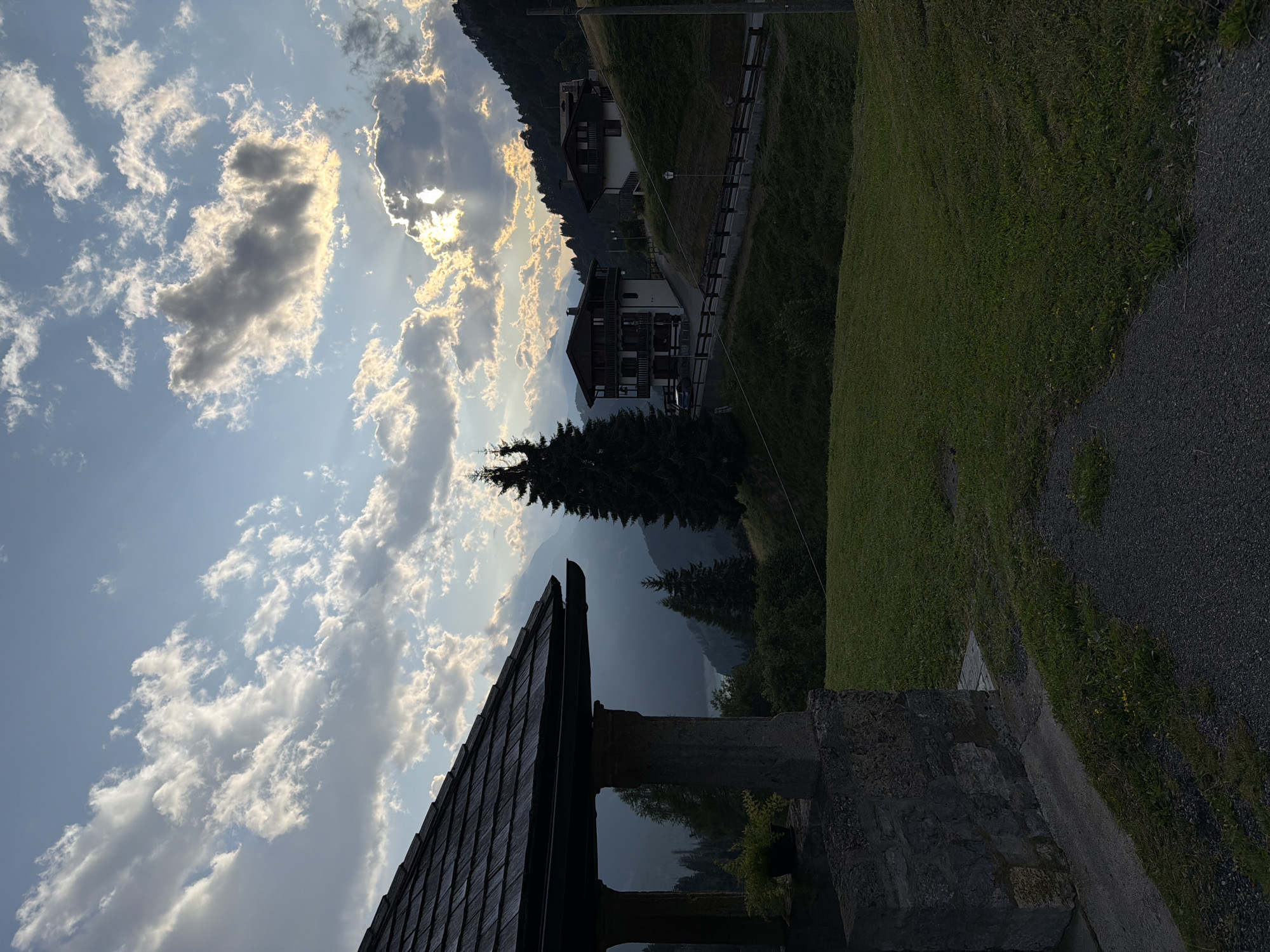 The image depicts a mountainous landscape in Sauris/Zahre, Italy, during what appears to be late afternoon or early evening. In the foreground, a stone structure with a dark wooden roof is visible on the left. Beyond this structure, a gently sloping grassy hill leads towards two multi-story houses built in a traditional alpine style. These houses are situated on the hillside, with a paved road and a wooden fence running between them and the foreground.
The background features rolling hills and mountains, partially obscured by mist or haze, suggesting a valley or a distant lake. The sky is dramatic, with a mix of light and dark clouds. Sunlight breaks through the clouds, casting rays of light and illuminating parts of the sky and the distant landscape. Several tall, dark evergreen trees are scattered throughout the scene, adding to the mountainous atmosphere. A car is parked on the road near the left-most house. The overall impression is one of a serene, rural mountain village at dusk.