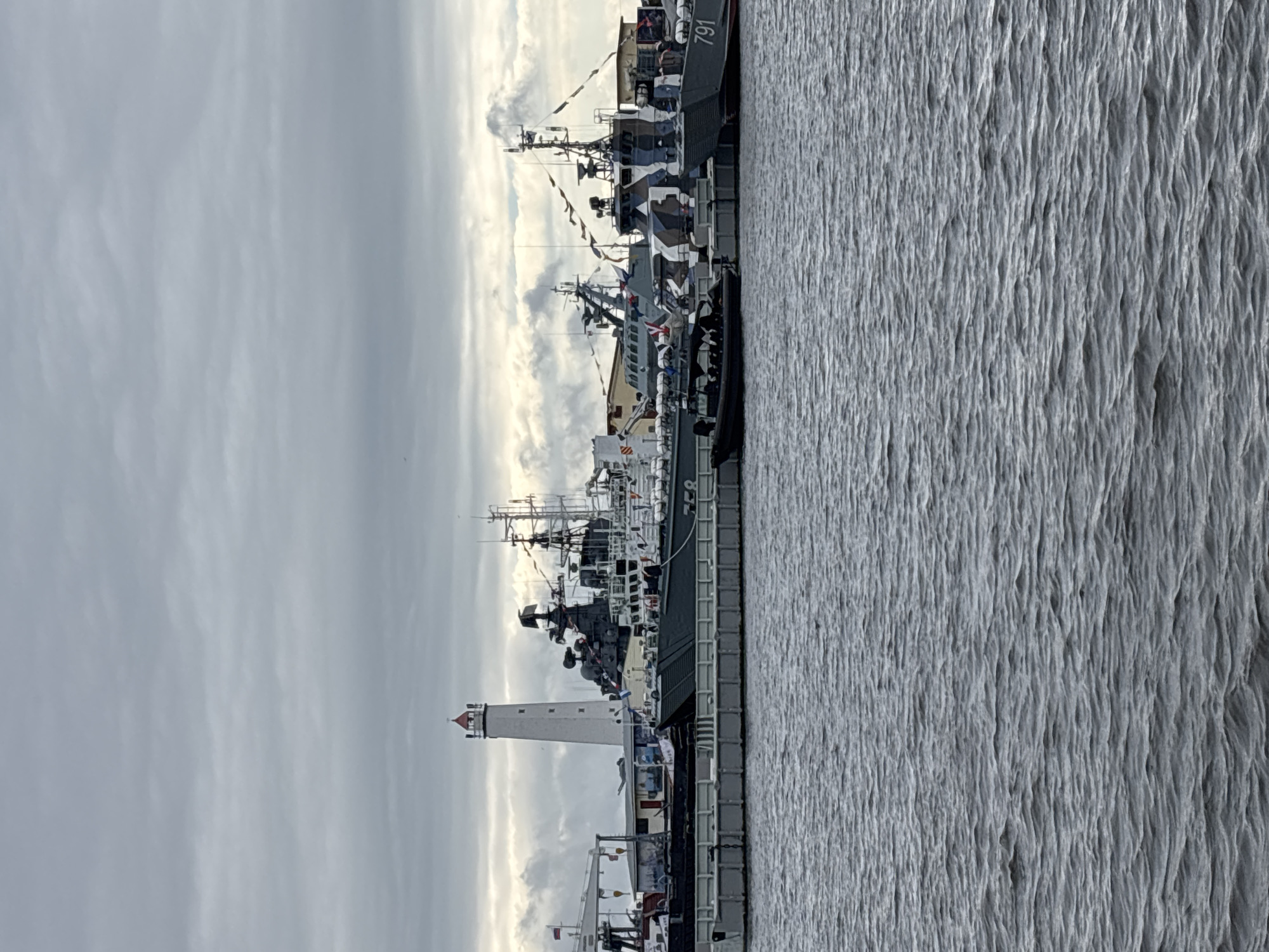 The image depicts a harbor scene in Kronstadt, Russia, on a cloudy day. In the foreground, choppy gray water fills the bottom half of the frame. Behind the water, a gray concrete pier runs horizontally across the image, with several vessels docked alongside it.
The most prominent vessel is a large, dark gray naval ship with a multi-level superstructure and antennas. It has the number "758" visible on its side. To the right of this ship is another naval vessel, also dark gray, with a distinctive camouflage pattern of black, white, and gray. This ship has the number "791" painted on its bow. A smaller, dark-colored rigid inflatable boat with several figures onboard is positioned in the water near the pier, in front of the larger ships.
In the background, a tall, white lighthouse with a red top stands out against the overcast sky. Several other buildings and structures, possibly part of the port infrastructure, are visible behind the ships and the lighthouse. Flags are seen flying from the ships and a pole near the lighthouse. The overall atmosphere is somber due to the cloudy weather.