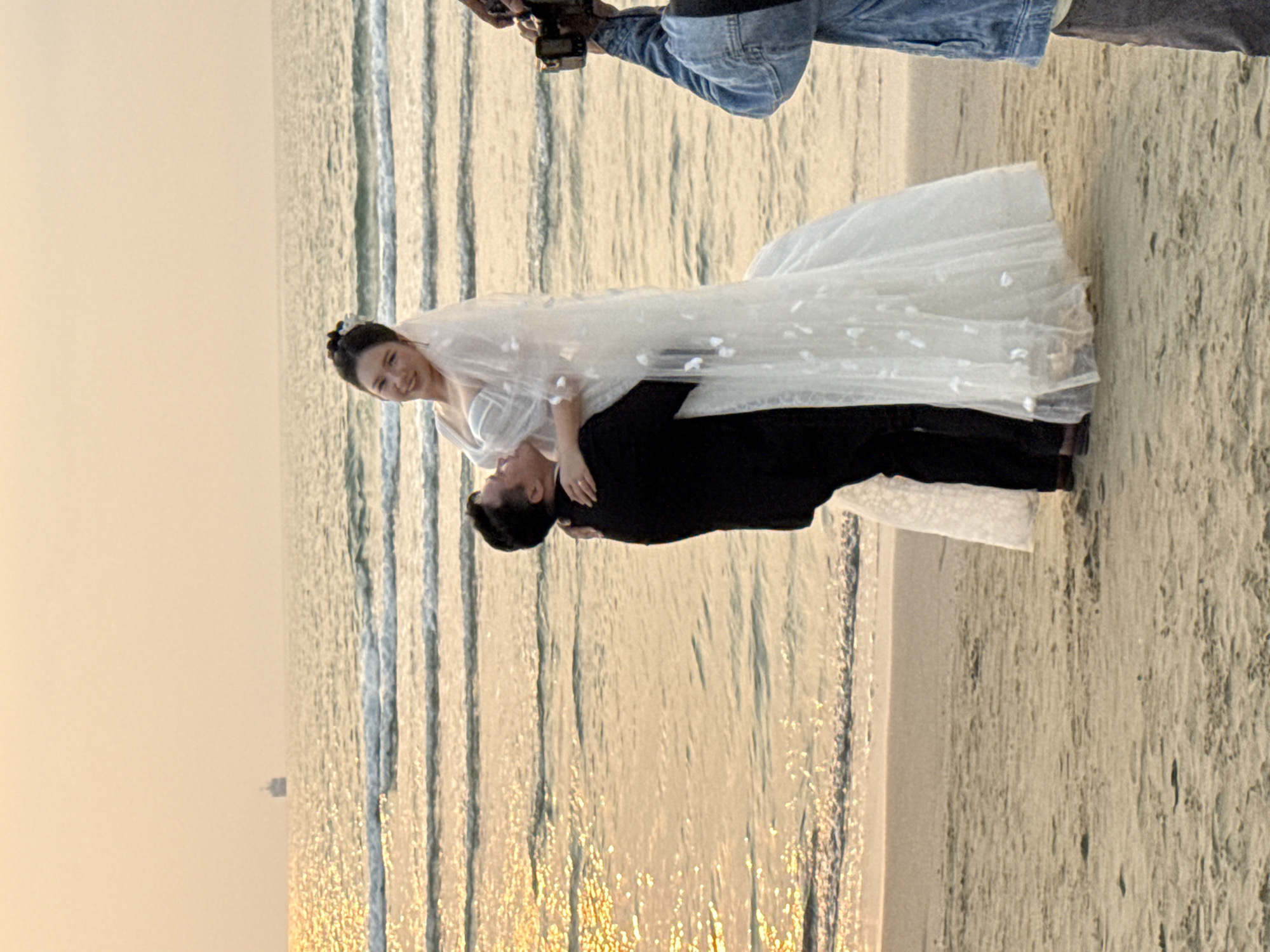 A wedding couple is being photographed on a beach in Đà Nẵng, Vietnam. The bride is wearing a white wedding dress with floral detailing, while the groom is wearing a black suit. He is holding her in a position where she is slightly leaning back and smiling. The beach setting includes the ocean with visible waves and a sandy shoreline. A person with a camera is present in the background, presumably taking the photo of the couple. The scene has a warm, golden hue, indicating that it is near sunrise or sunset.