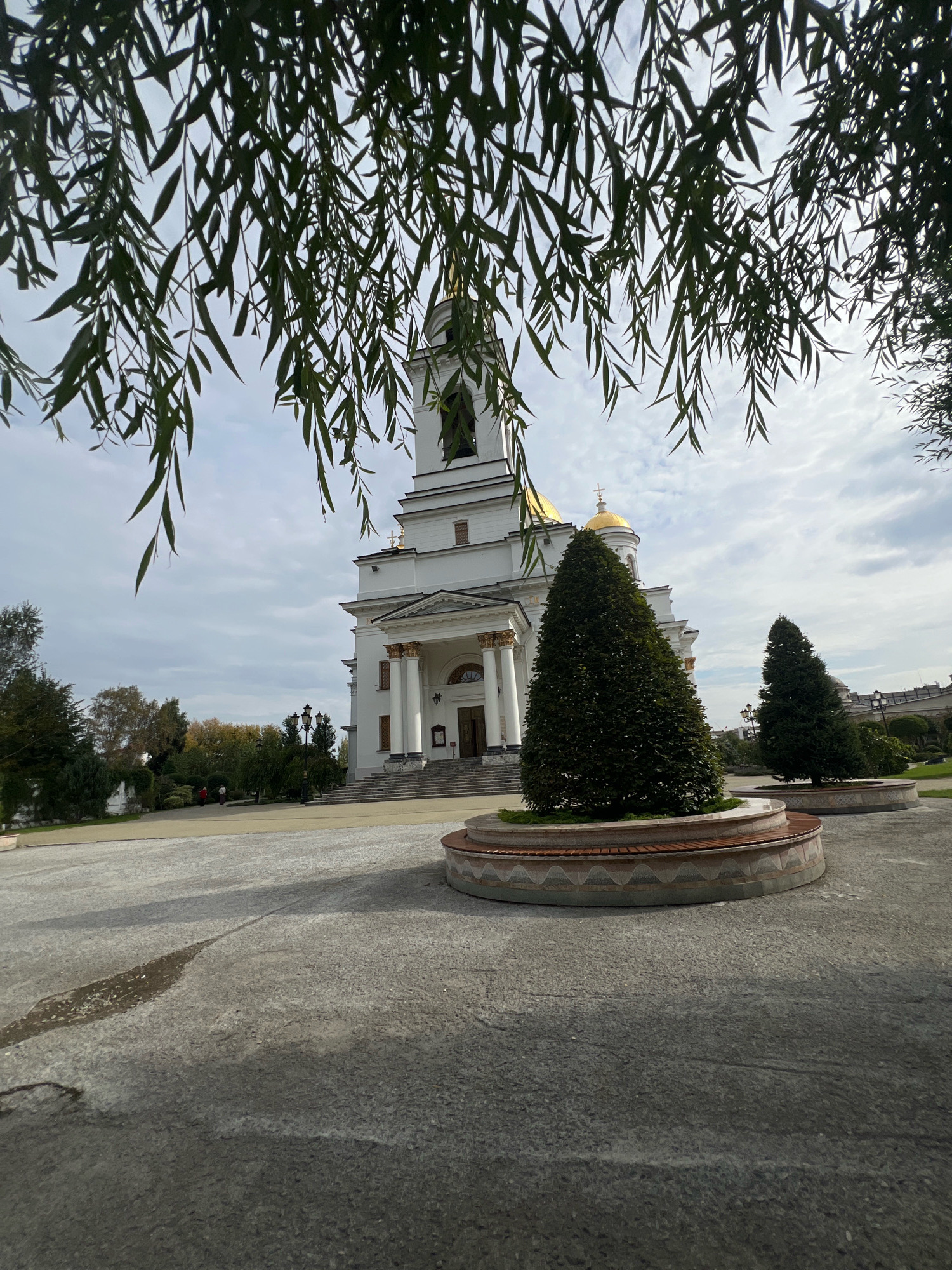 The image captures the Church of All Saints in Yekaterinburg, Russia, viewed from a courtyard. The church is a grand white building with a prominent bell tower topped with a cross and a golden dome. Its facade features classical architectural elements, including columns and a grand staircase leading to the entrance. In the foreground, a circular planter with a sculpted evergreen tree is prominently featured, with a curved bench surrounding its base. Another smaller evergreen tree is visible further back, also within a planter. The courtyard itself is paved with grey stone, and a wet patch suggests recent rain. Overhanging the scene from the top are the long, drooping branches of a willow tree, framing the view of the church. The sky is overcast with scattered clouds. Two individuals are visible in the middle ground, walking across the courtyard, and a few more are in the distance near the trees.