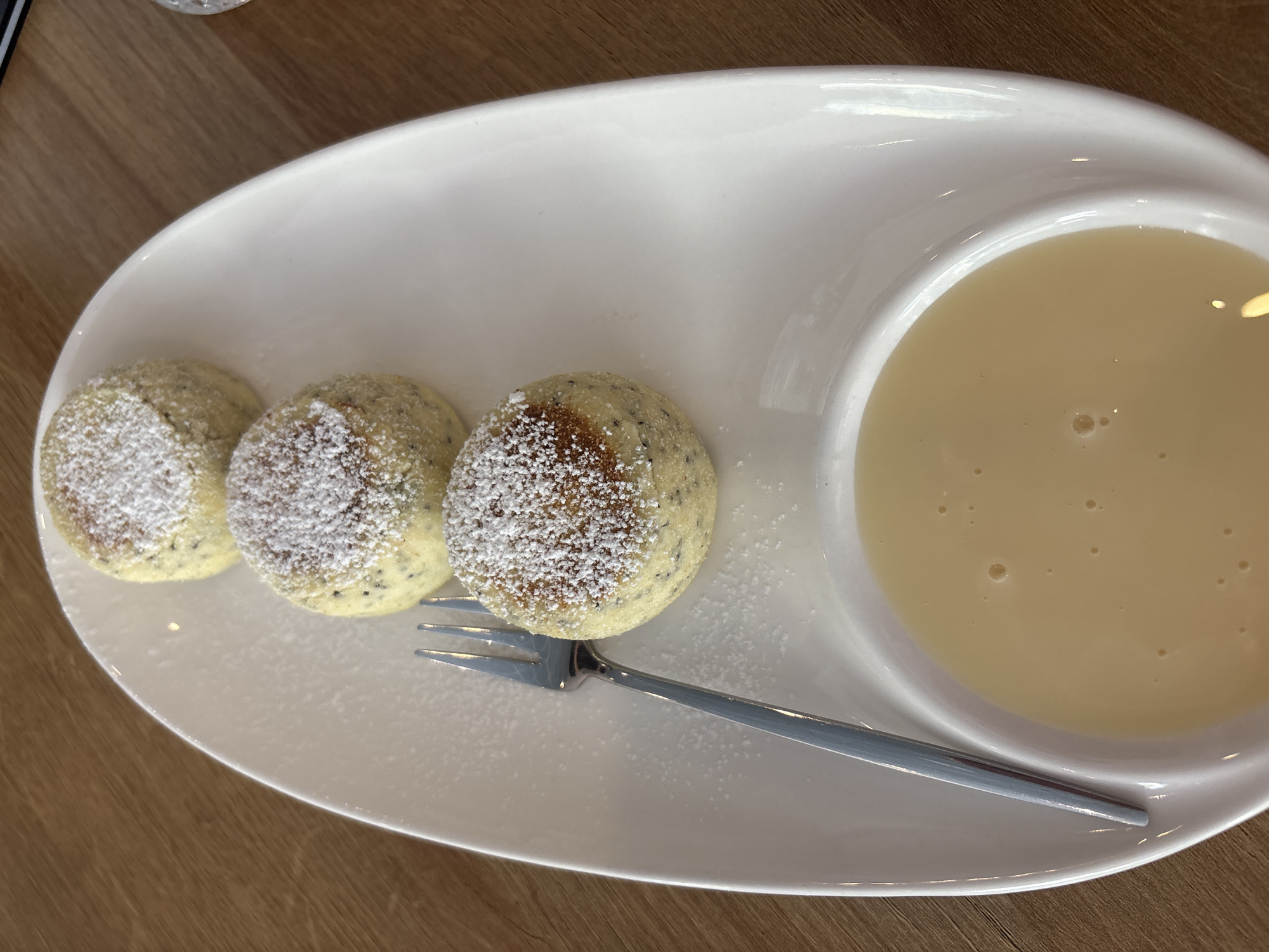 A top-down shot depicts a white, oval-shaped plate containing three small, round, cake-like items dusted with powdered sugar, arranged linearly. A small fork with a silver-colored handle lies adjacent to the cakes. A circular depression on the right side of the plate contains a light-colored, creamy liquid, likely a sauce or custard. The plate rests on a wooden surface. The location is unidentified, and no people or actions are visible. There is no way to confirm the location is Bắc Nha Trang, Vietnam.