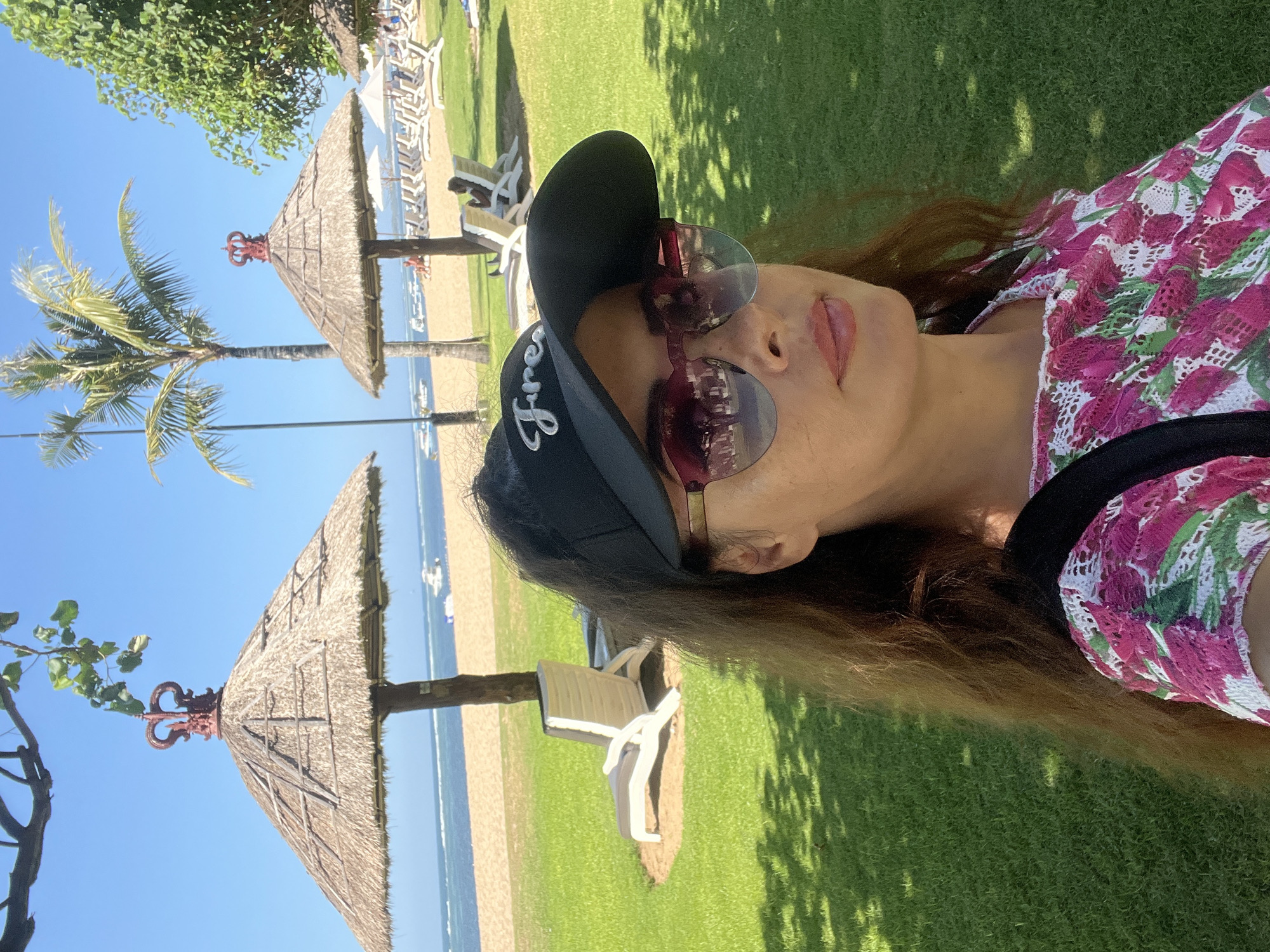 A medium shot captures a woman with long, wavy brown hair taking a selfie on a sunny day at a beach resort in Kuta Selatan, Indonesia. She is wearing a black visor with the word "Frees" embroidered in white, stylish pink-rimmed sunglasses that reflect the scene, and a pink and white floral print top. She is positioned in the foreground, with a lush green lawn behind her. In the mid-ground, there are several white lounge chairs and thatched-roof umbrellas on the sand, with a calm blue ocean and a few boats visible in the background. A palm tree and another tree with green foliage are also present. The sky is clear and blue.