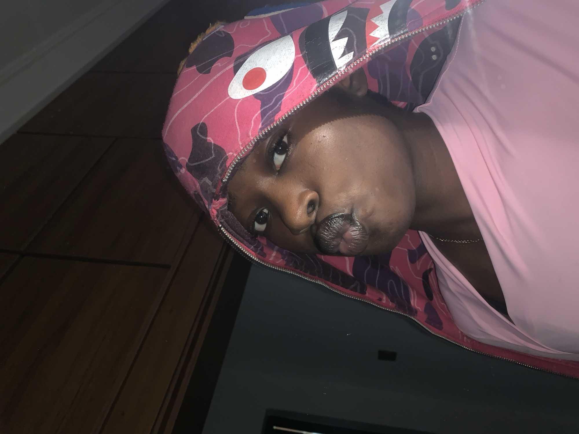 In Gura Top, Nigeria, a young person with dark skin, wearing a pink hooded jacket with a camouflage pattern and a shark design, is captured in a close-up portrait. The person's lips are pursed in a pout, and their eyes are looking directly at the camera. They are also wearing a light pink top and a delicate gold chain. The background consists of wooden paneling and a dark wall, suggesting an indoor setting, possibly a home.
